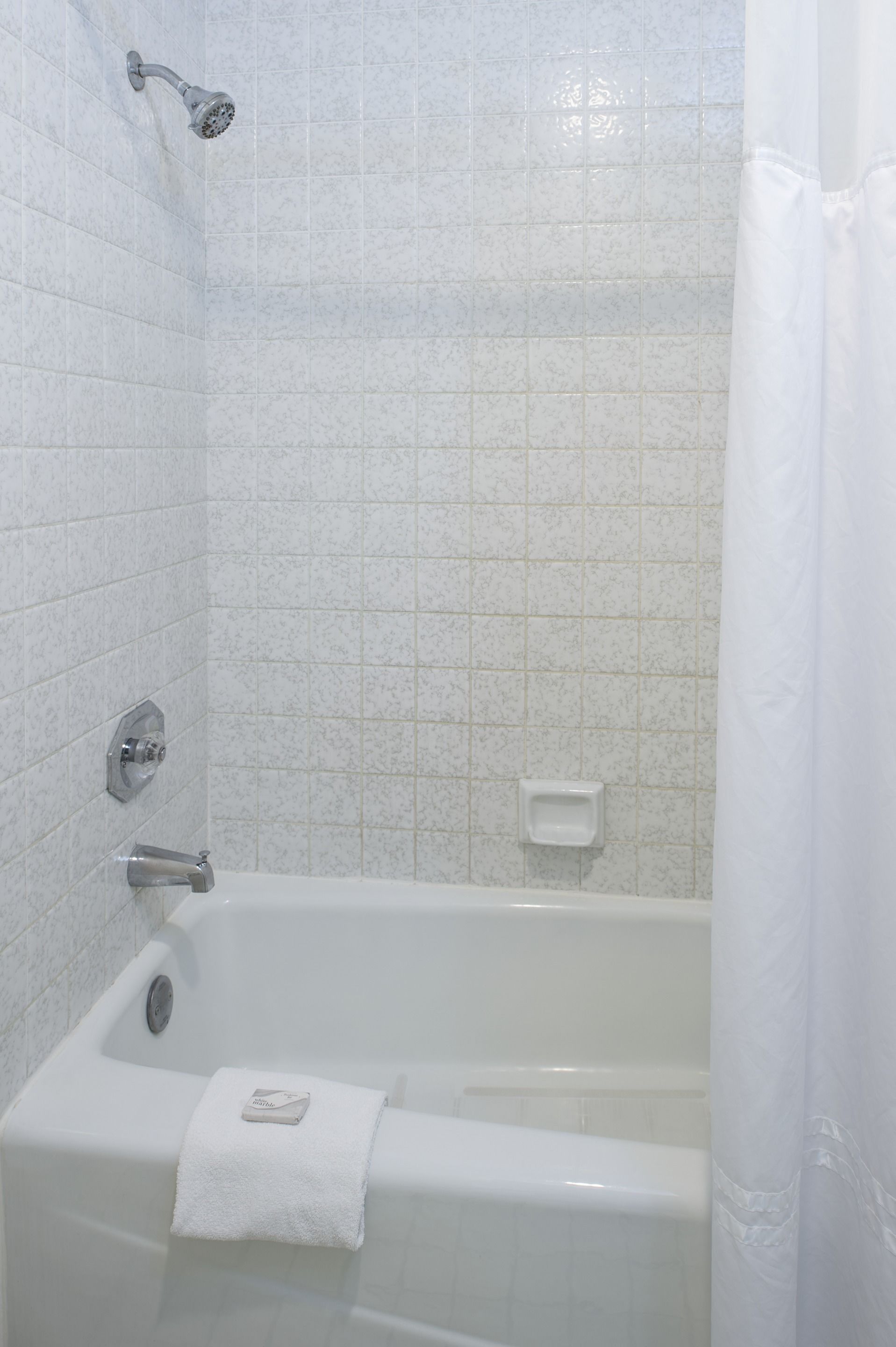 Une salle de bain aux murs recouverts de carreaux blancs, avec une baignoire, une pomme de douche et un porte-savon.