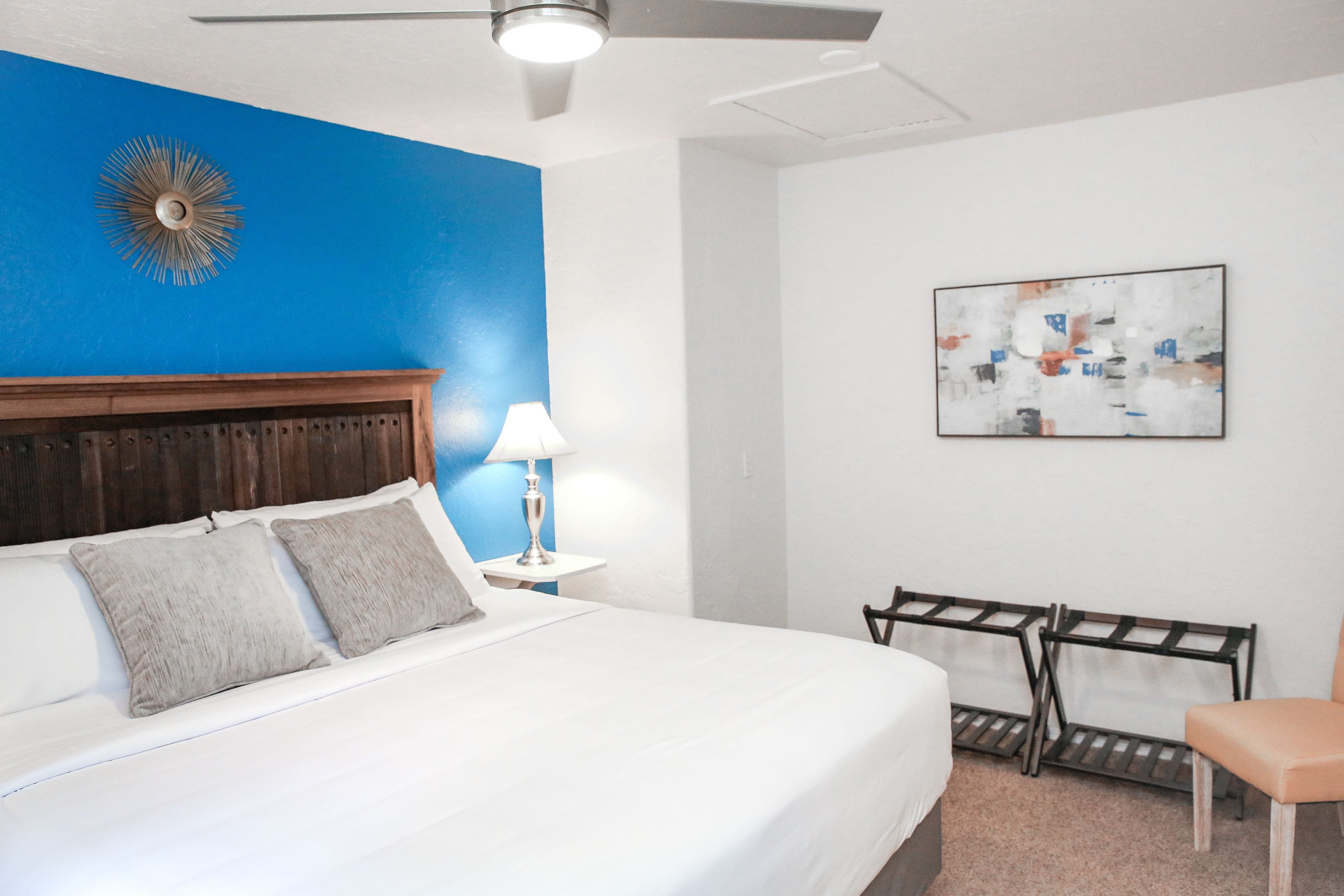 Habitación de hotel con cama, mesita de noche, lámpara, silla, una obra de arte en la pared y ventilador de techo.