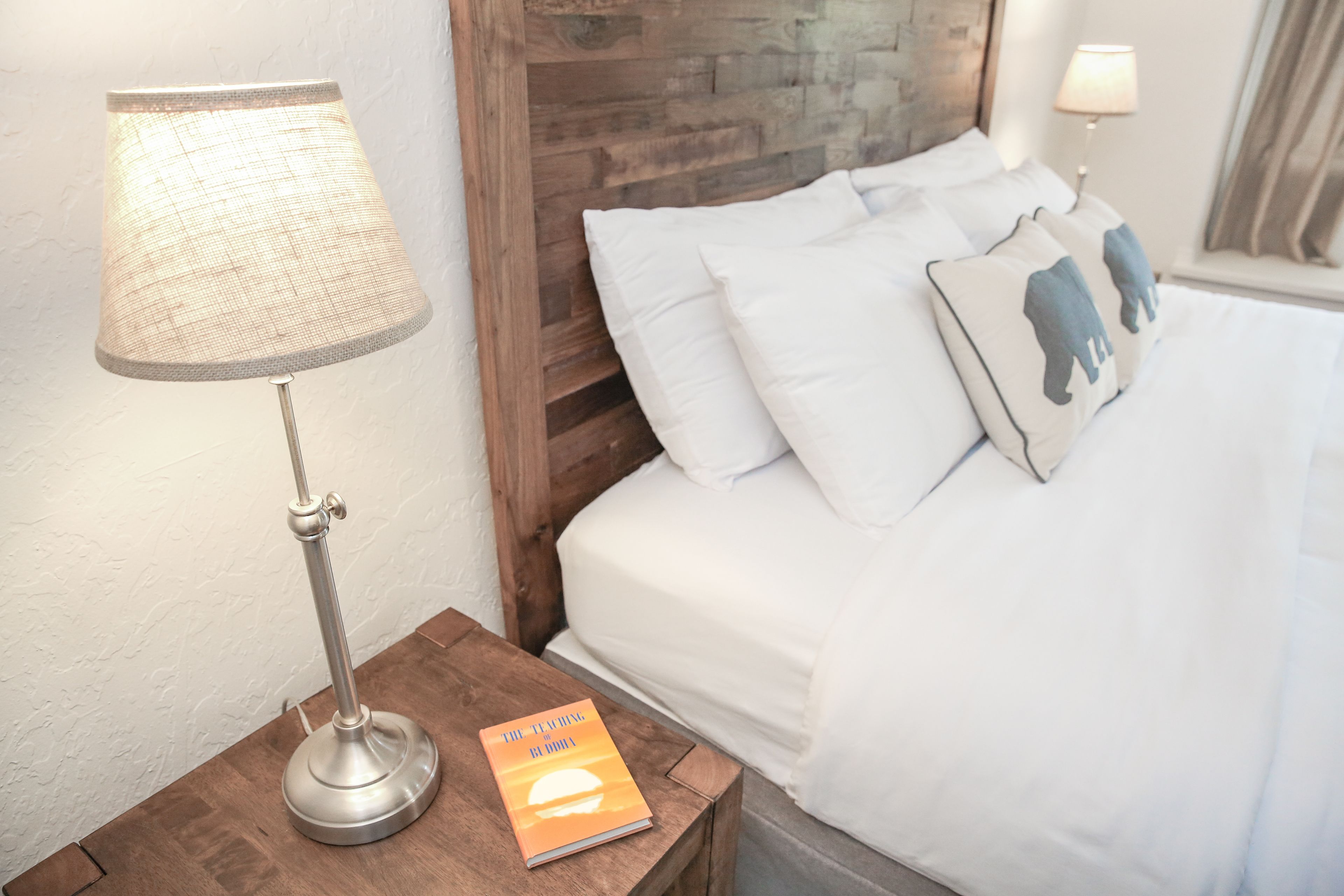 Ein ordentlich bezogenes Bett mit weißen Leinen und dekorativen Kissen, eine Nachttischlampe und ein Buch auf einem Holznachttisch.