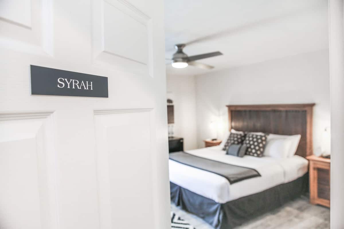 A bedroom with a bed, nightstands, and a ceiling fan. A sign on the door reads "SYRAH".