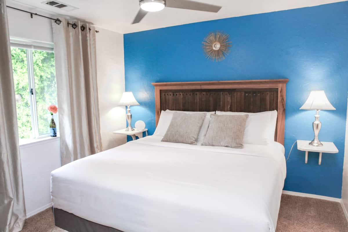 A bedroom with a large bed, two bedside tables with lamps, a ceiling fan, and a window with curtains.
