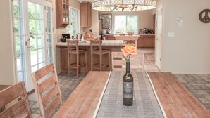 Een eetgedeelte met een houten tafel, stoelen, een vaas met een bloem en een fles wijn.