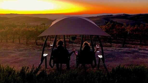 Twee personen zitten onder een grote parasol in een wijngaard bij zonsondergang.