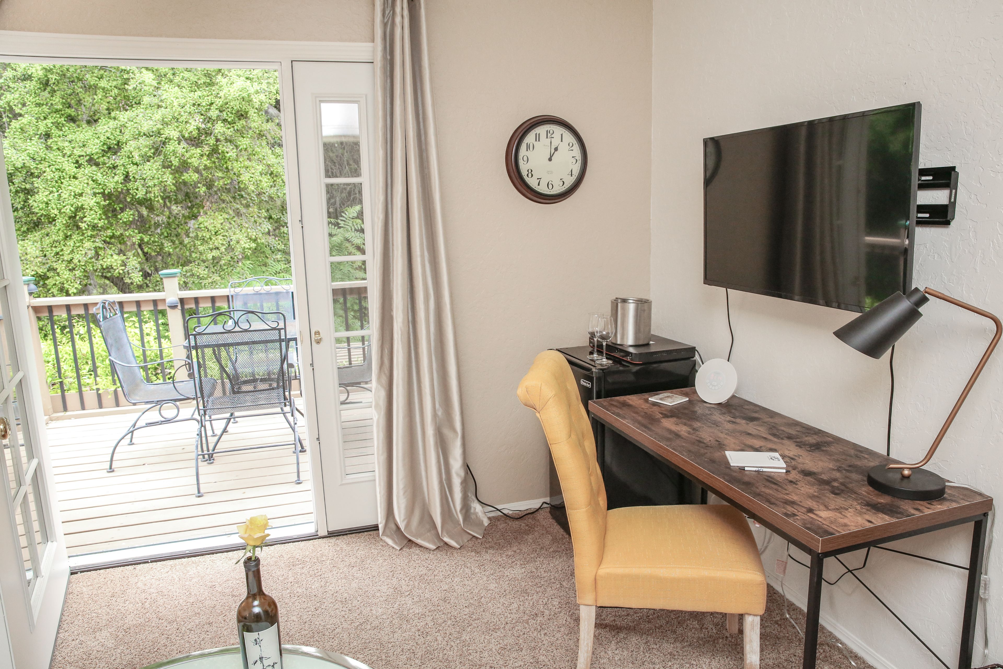 Una habitación con escritorio, silla, televisor y vistas a un balcón con mobiliario exterior.