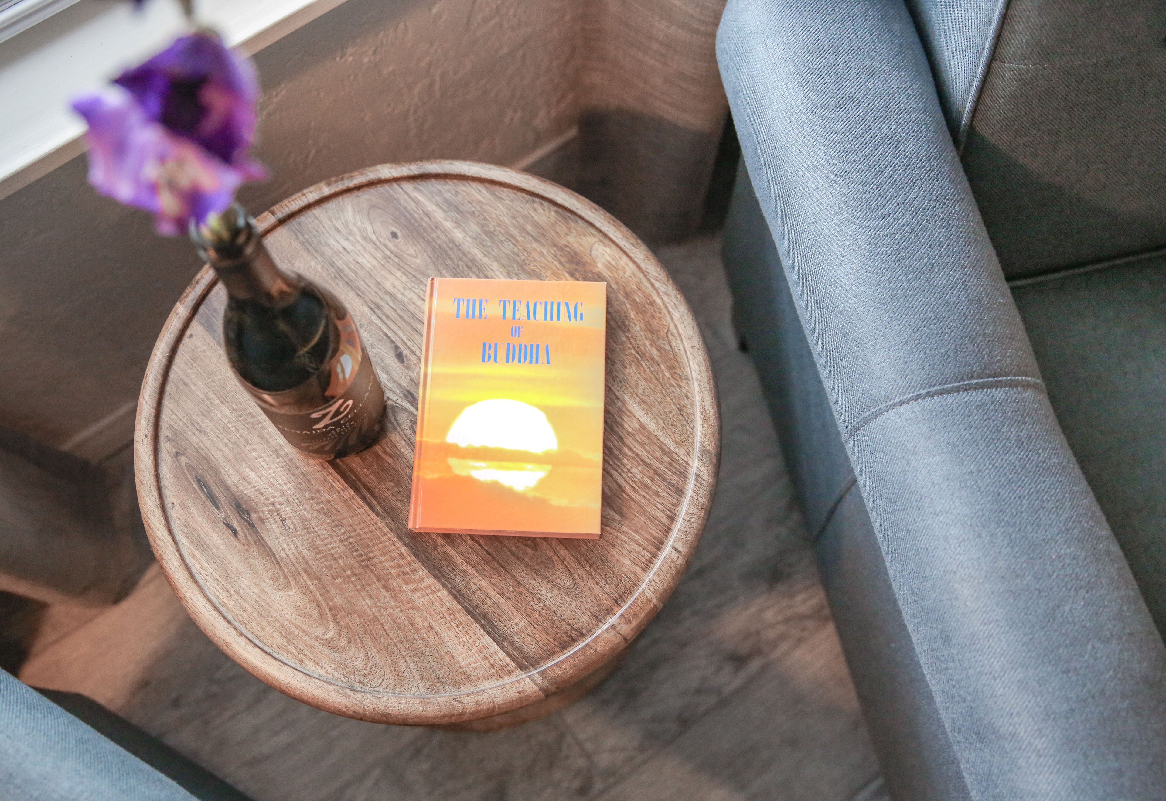 A book titled "The Teaching of Buddha" on a wooden side table with a vase of purple flowers.