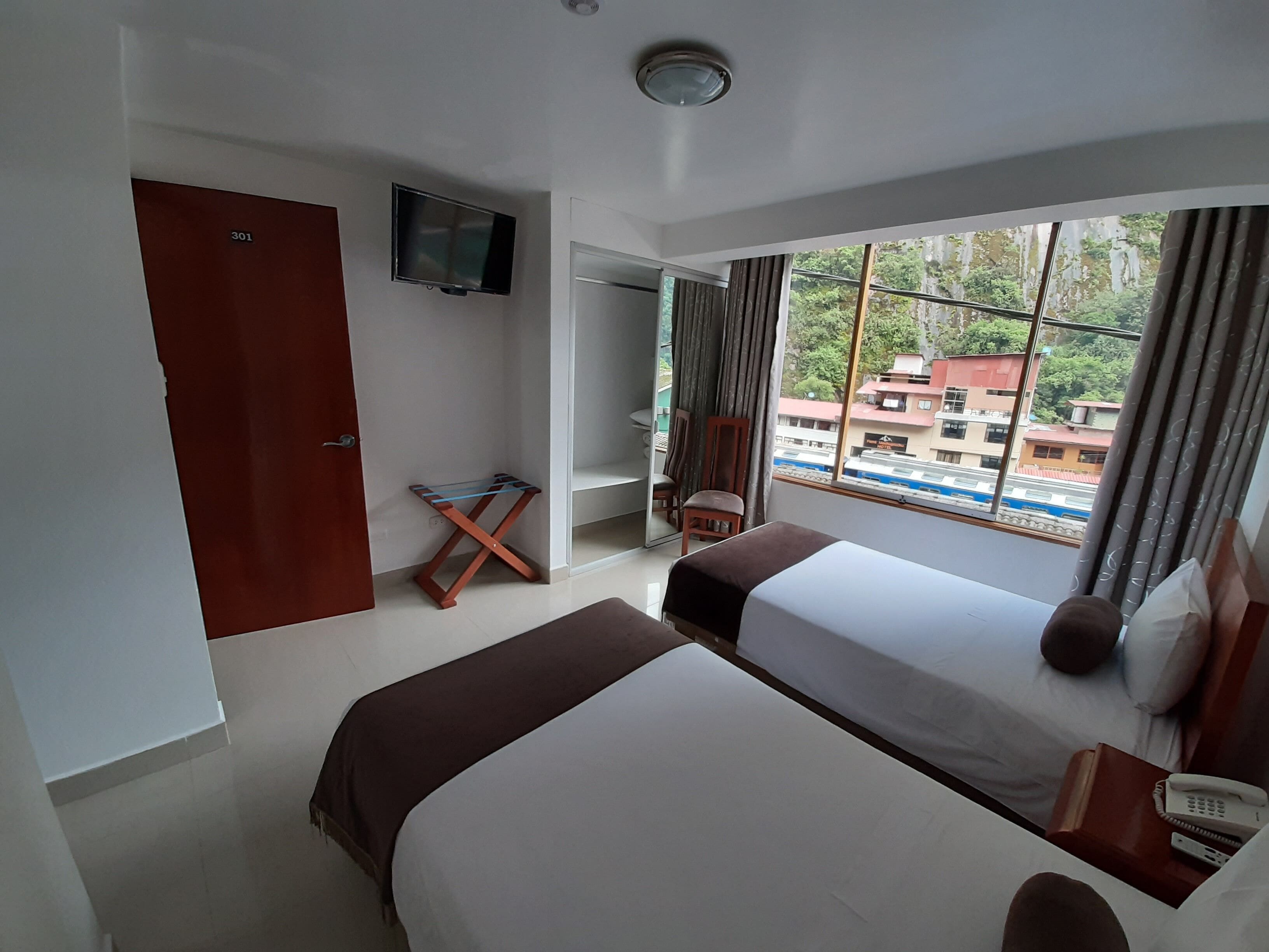 A hotel room with two beds, a TV mounted on the wall, a small table, and a window with a view of buildings and a train.