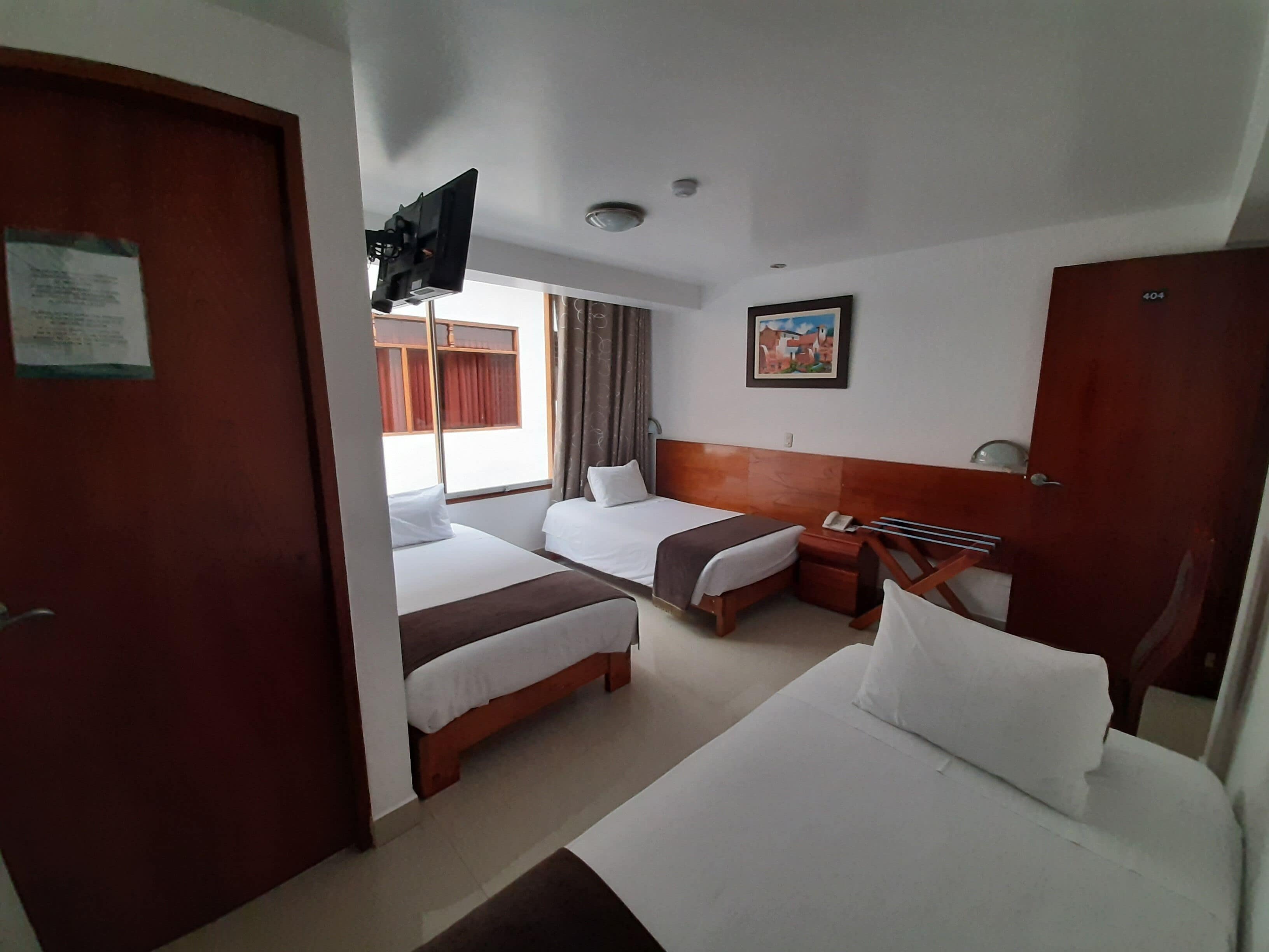 A hotel room with two beds, a wooden headboard, a flat-screen TV mounted on the wall, a painting above the headboard, and a door with a sign.