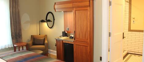 A hotel room with a bed, a desk with a computer, a chair, a wardrobe, and a bathroom visible through an open door.
