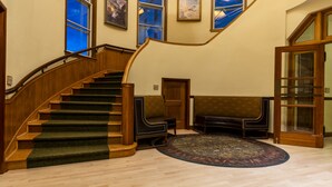 A grand staircase with wooden railings and carpeted steps, leading to a landing with a large, ornate rug and a seating area with leather sofas.