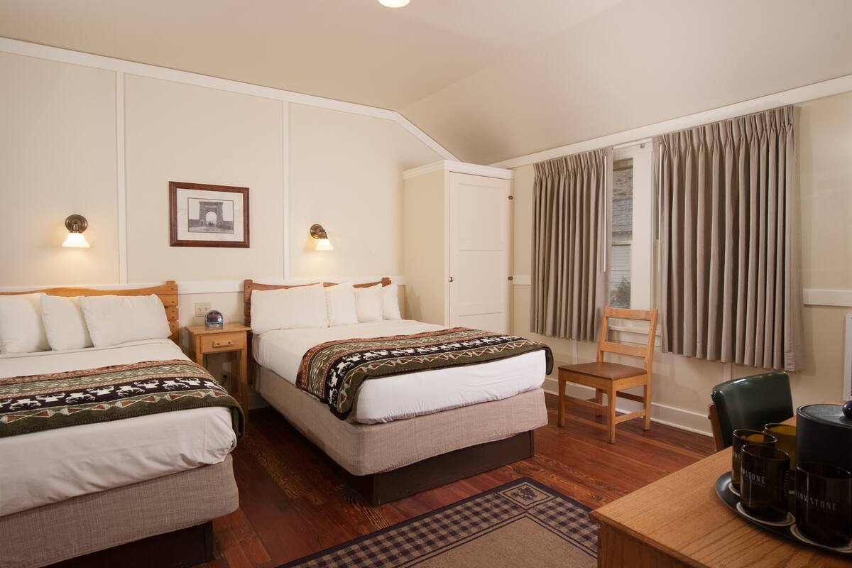Ein Hotelzimmer mit zwei Betten, einem Holzstuhl, einem Tisch mit Tassen und Blick durch Vorhänge nach draußen.