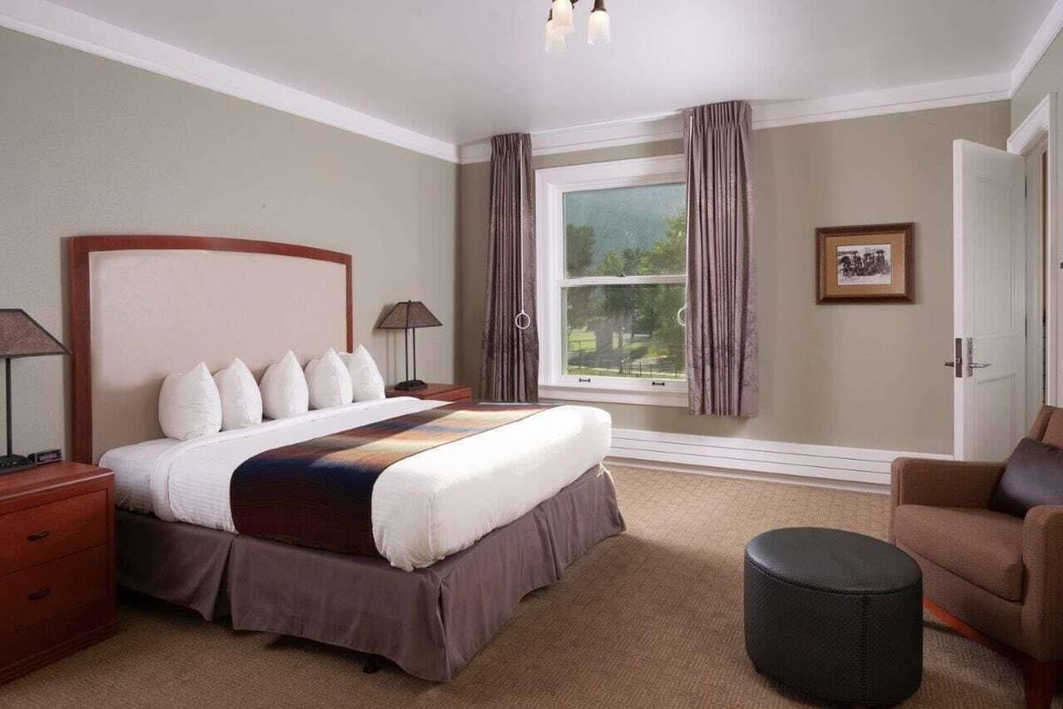Ein Hotelzimmer mit einem großen Bett, einem Sessel, einem runden Hocker, einem Nachttisch, einer Lampe und einem Fenster mit Vorhängen.