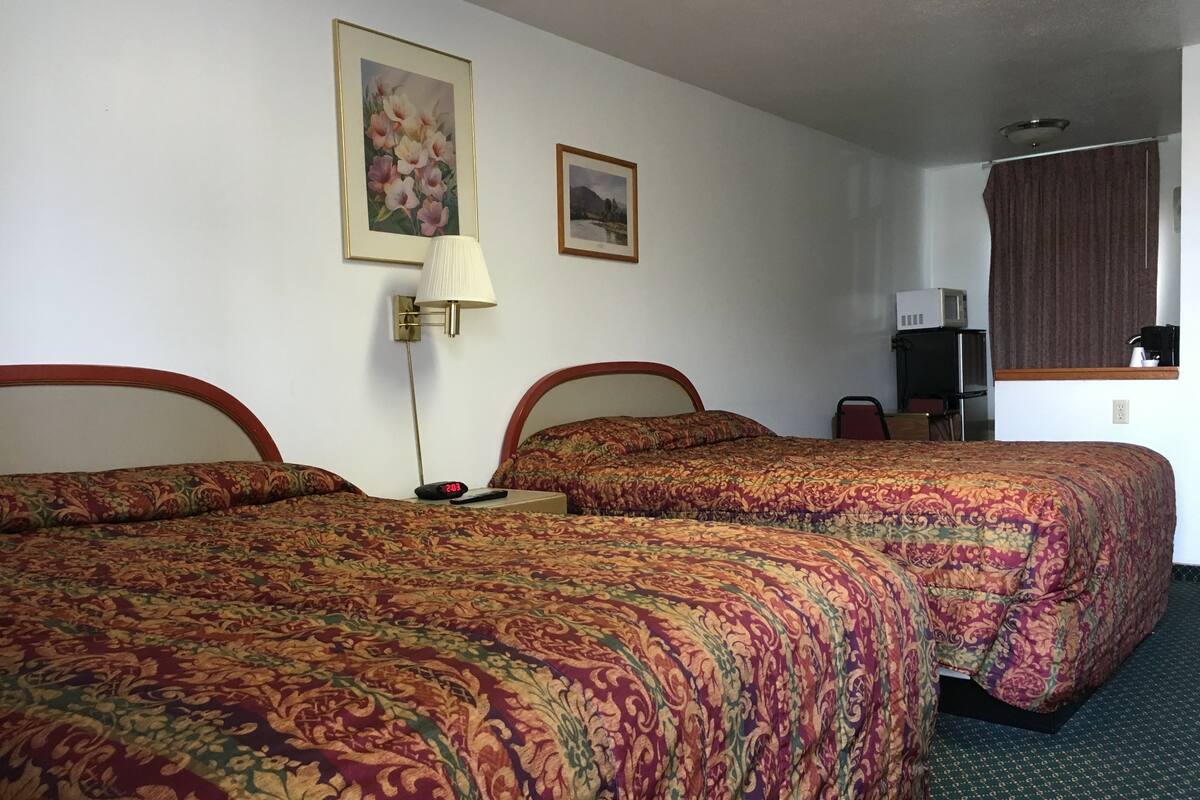 A hotel room with two beds, a nightstand with a lamp, a small table with a coffee maker, and a picture on the wall.