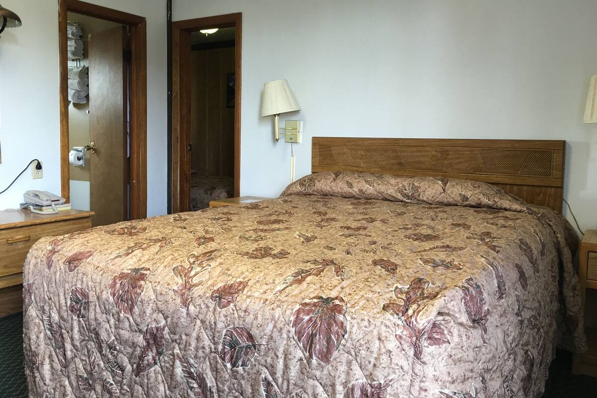 A hotel room with a large bed, a wooden headboard, a bedside lamp, a nightstand, and a door leading to another room.