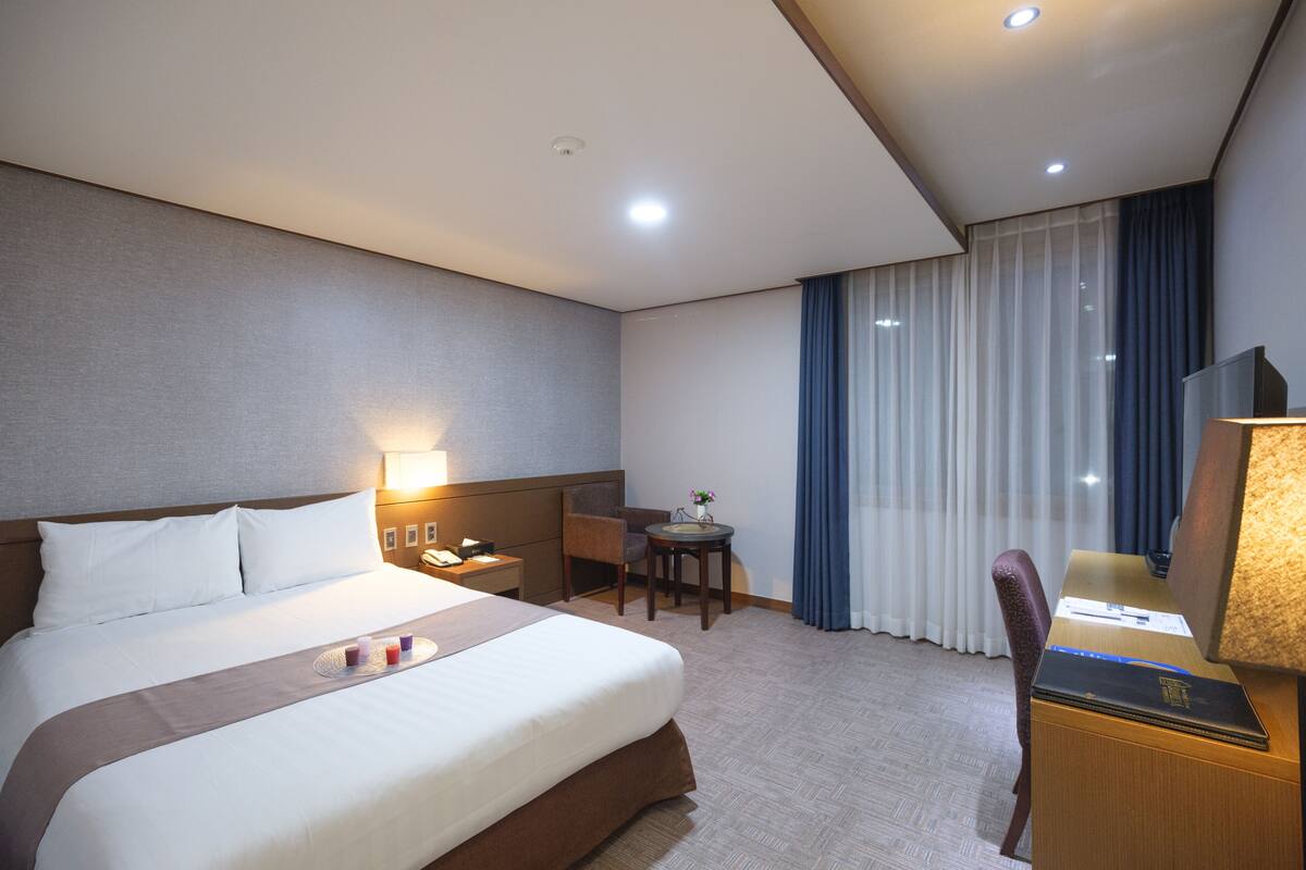 Una habitación de hotel con una cama grande, un escritorio con lámpara, una silla, una mesita con un florero y una ventana con cortinas.