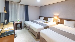 Une chambre d’hôtel avec trois lits, un bureau avec un ordinateur, une petite table avec une lampe et une fenêtre avec des rideaux.
