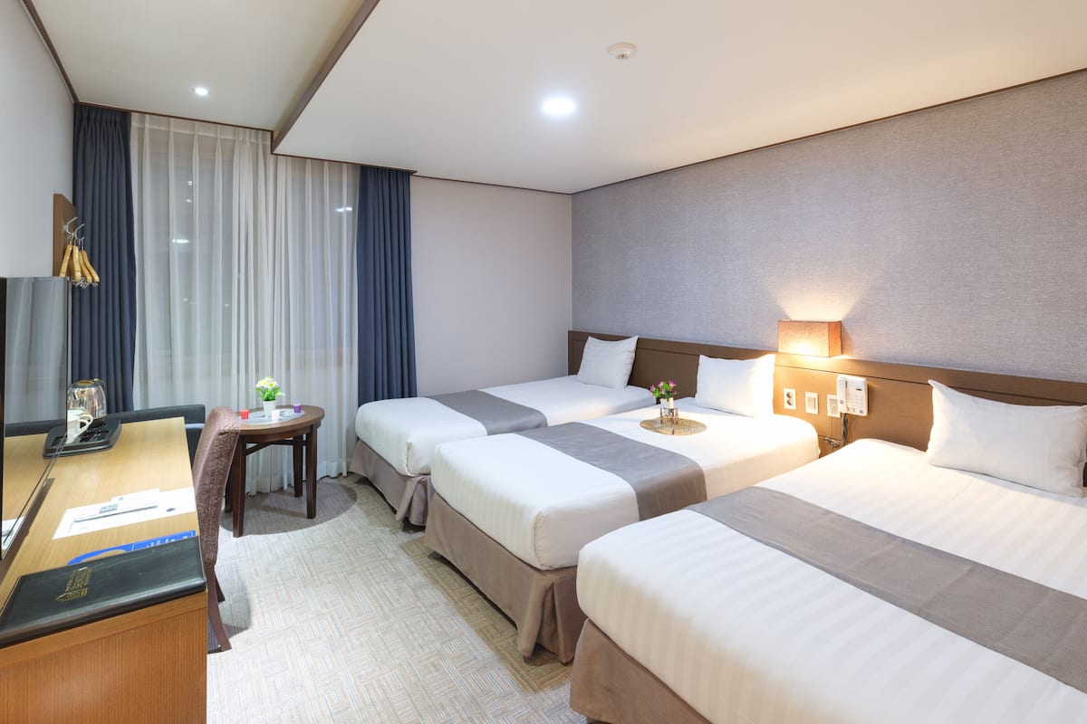 Habitación de hotel con tres camas, un escritorio con ordenador, una mesita con lámpara y una ventana con cortinas.