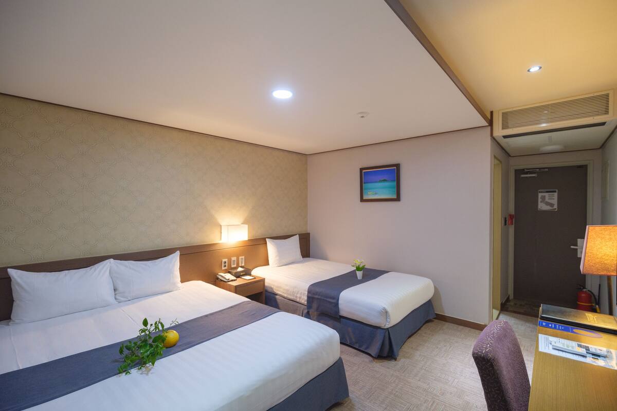 Habitación de hotel con dos camas, un escritorio, una silla, una mesita de noche y un aire acondicionado instalado en la pared.