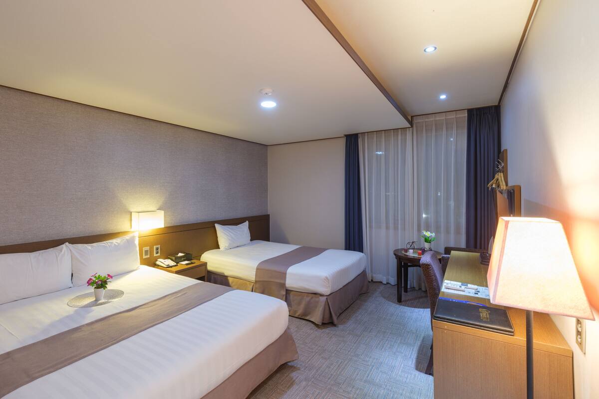 Habitación de hotel con dos camas, un escritorio, una lámpara, una silla y una mesita con un florero.
