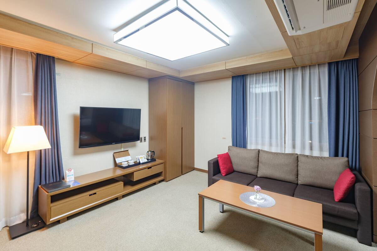 Una habitación de hotel moderna con televisión de pantalla plana, un sofá con cojines rojos, una mesita de madera y una lámpara de techo.