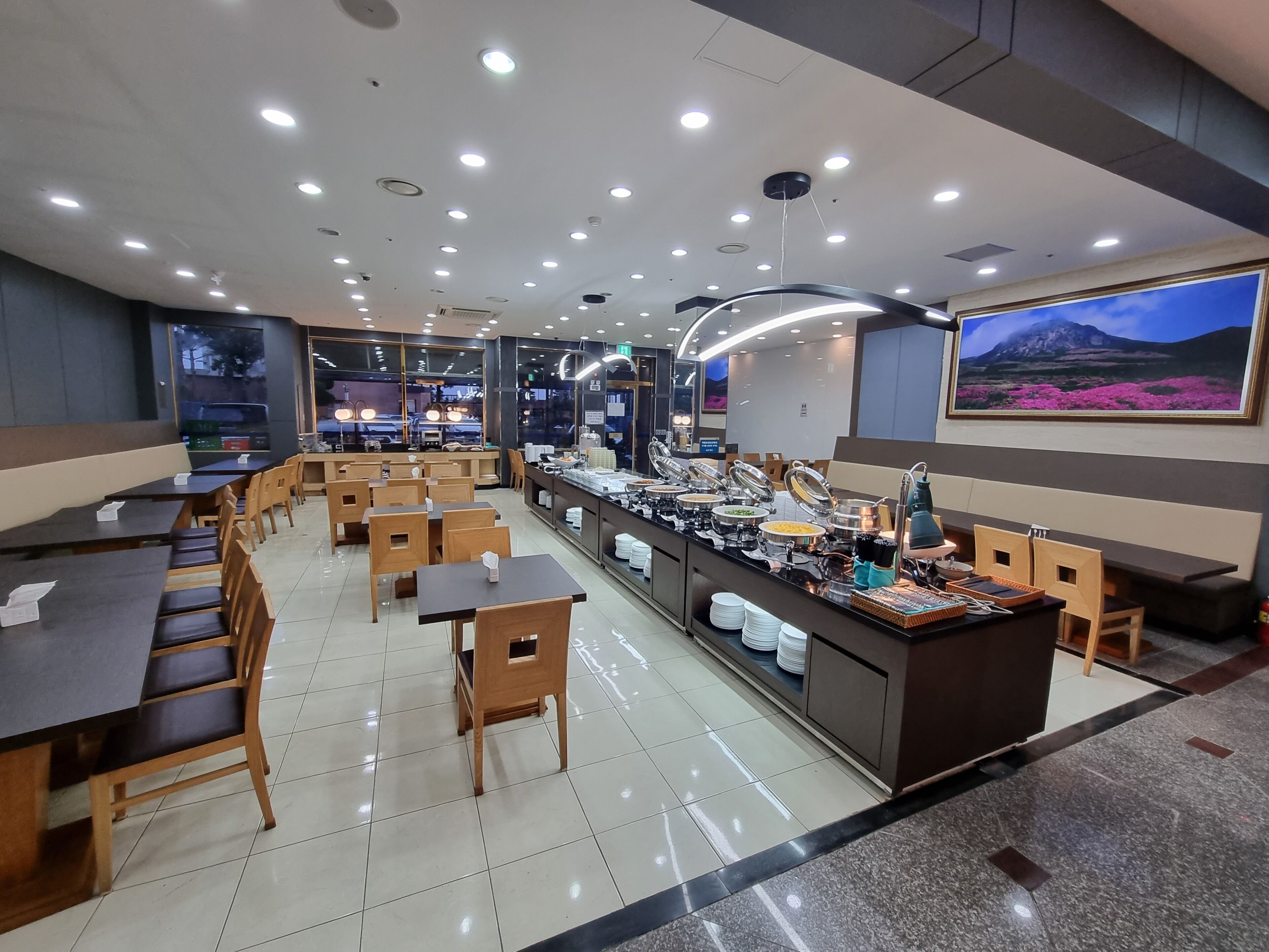Un restaurant doté de nombreuses tables et chaises, d’un buffet proposant une variété de plats, et d’un grand tableau encadré accroché au mur.