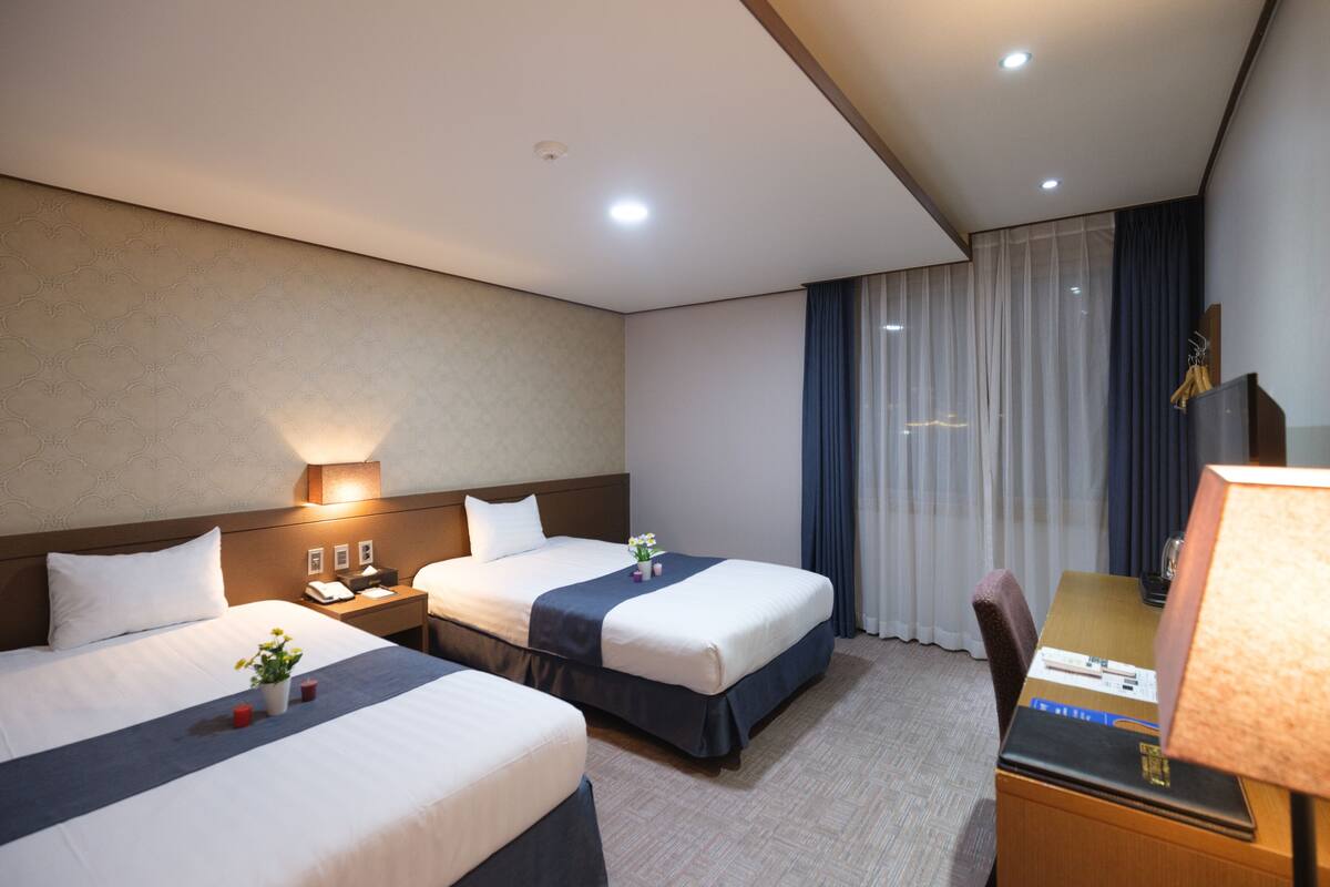 Habitación de hotel con dos camas, un escritorio, una silla, una lámpara y un ventanal con cortinas.
