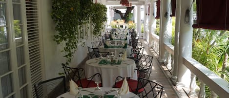 A covered outdoor dining area with round tables set for two, white columns, and green plants.