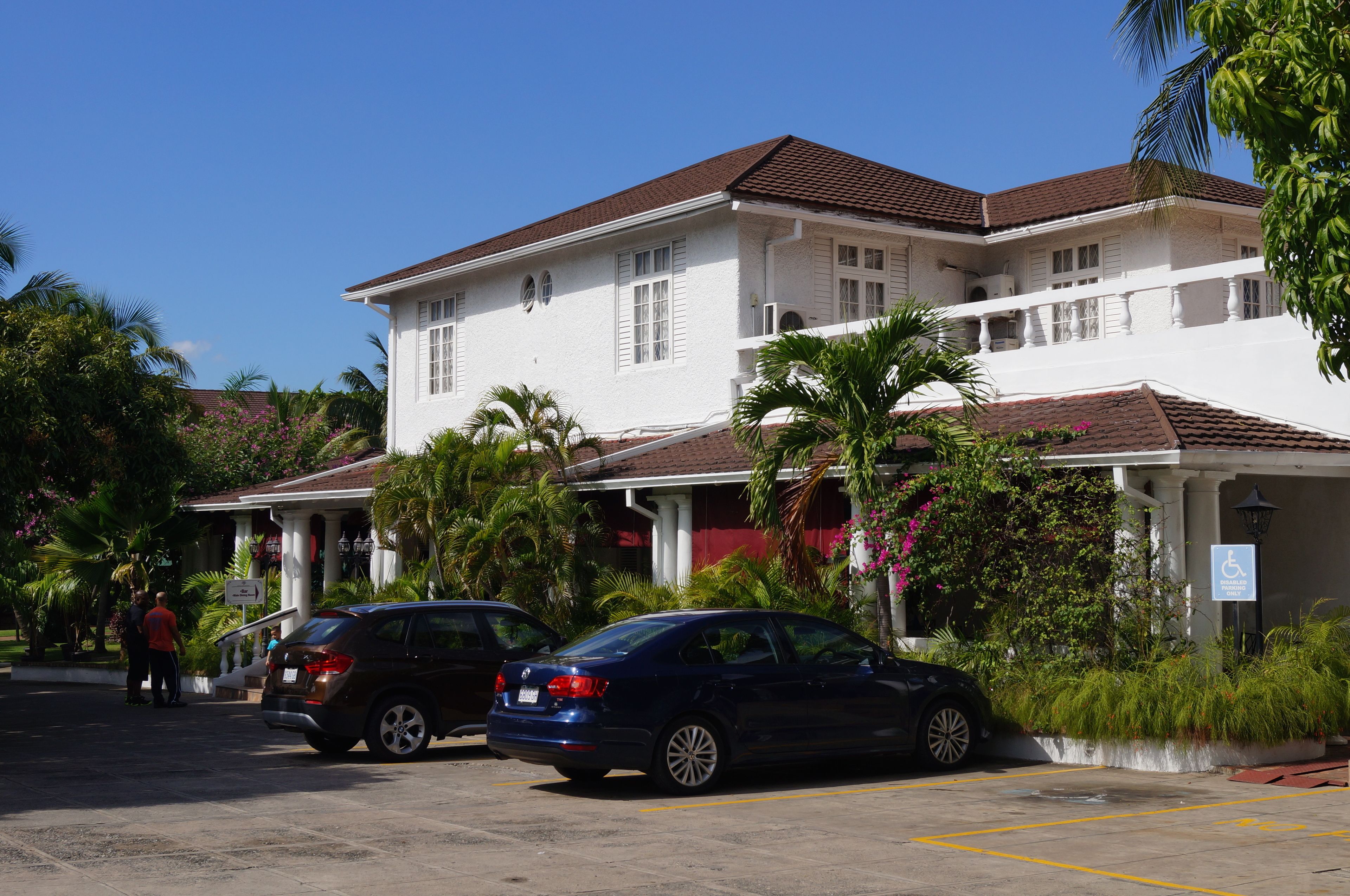 A two-story building with a white facade and a red-tiled roof, surrounded by lush greenery and palm trees.