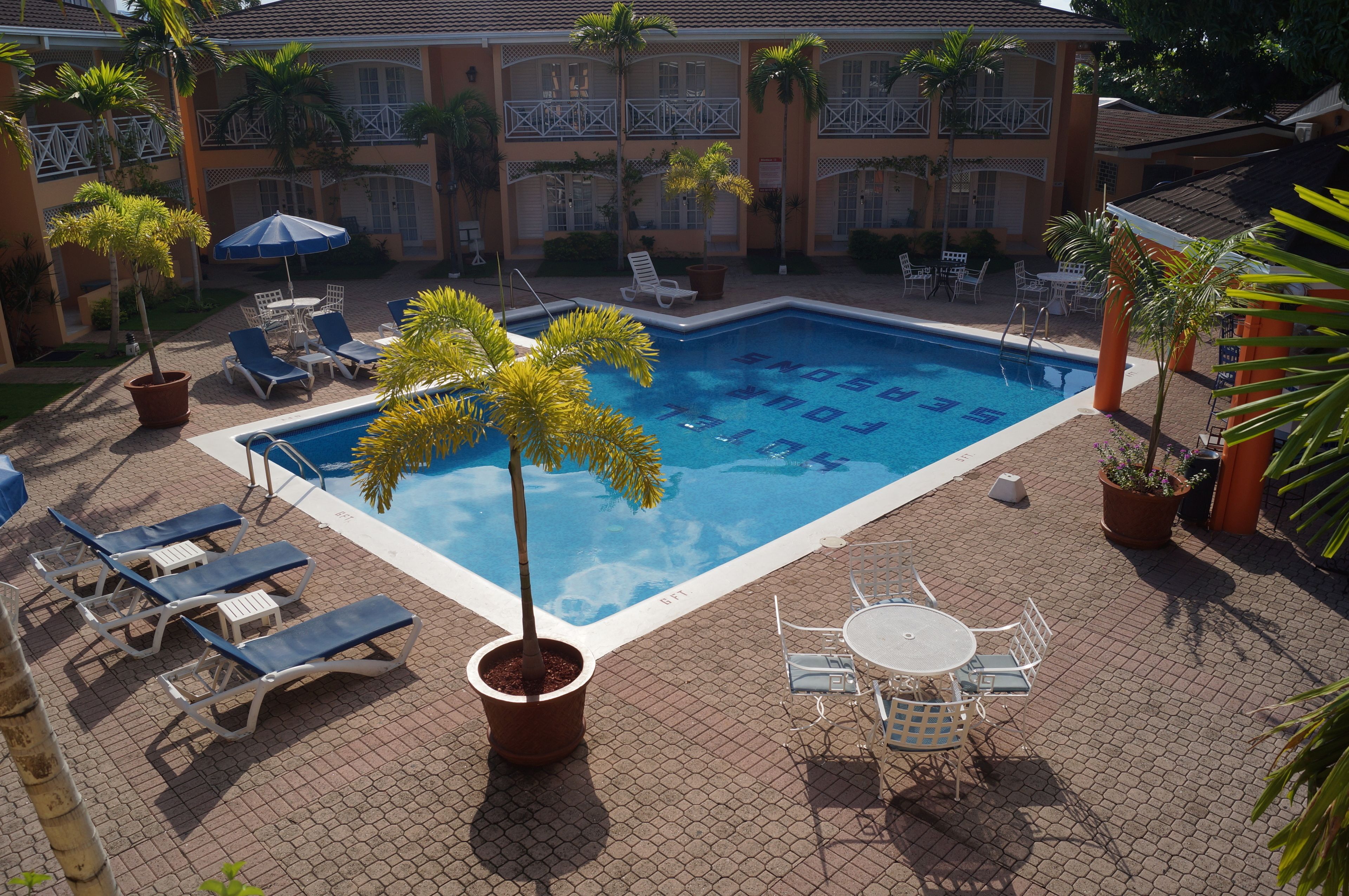 A swimming pool with lounge chairs and tables, surrounded by potted plants and a building with balconies.
