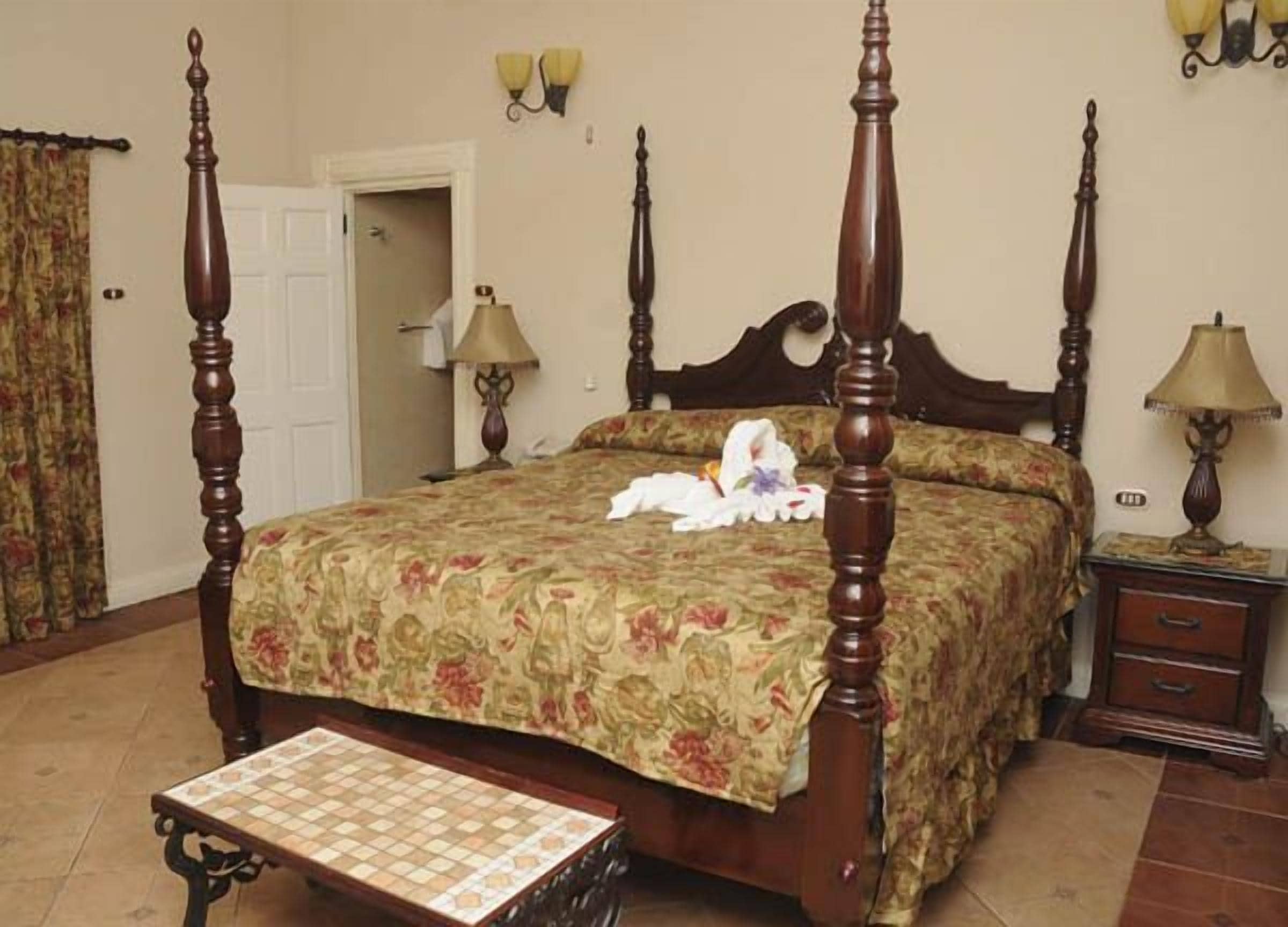 A four-poster bed with floral bedspread, a nightstand with a lamp, and a small table with a checkered tablecloth.