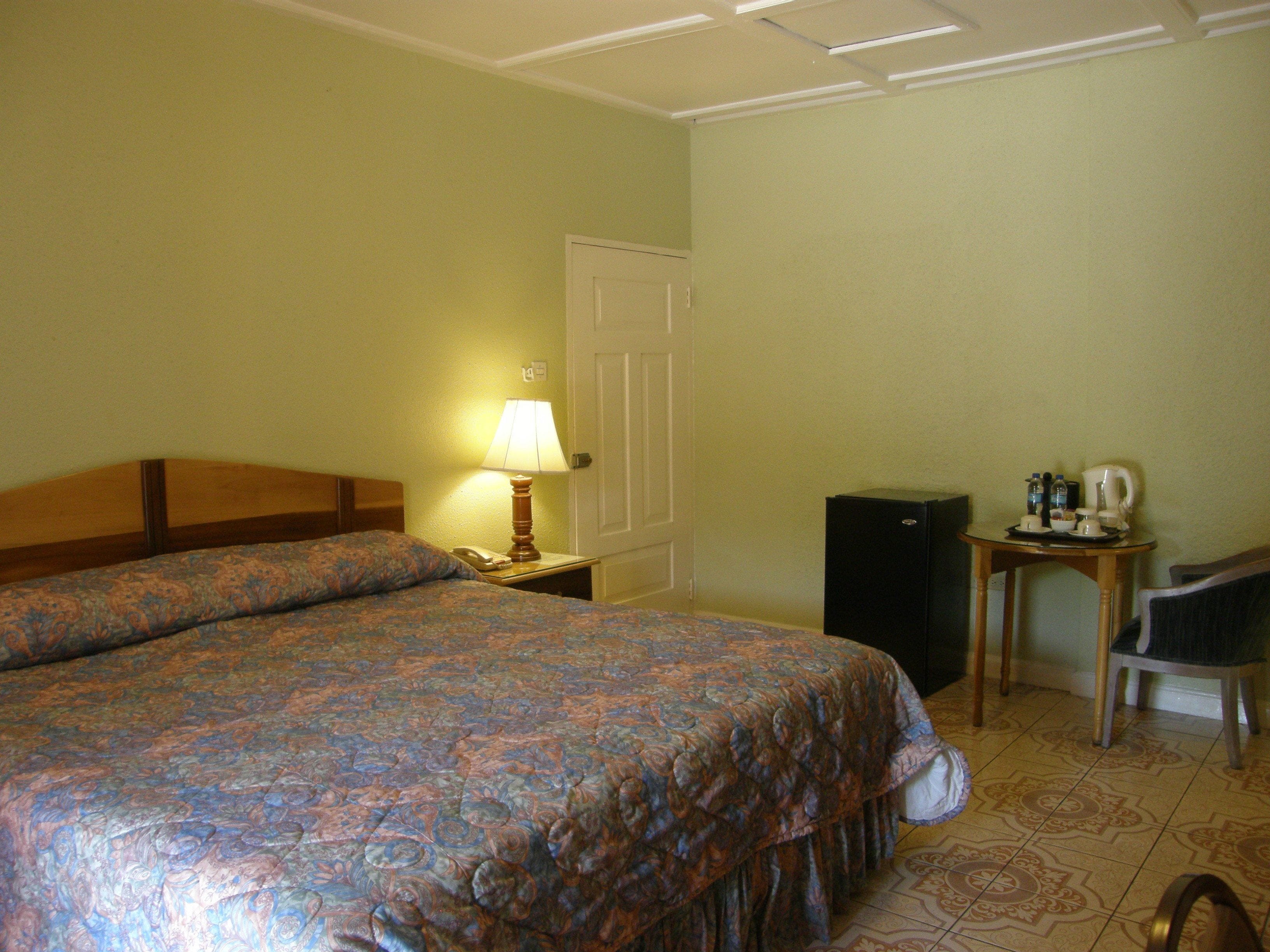 A hotel room with a large bed, a bedside table with a lamp, a small table with a coffee maker, and a chair.
