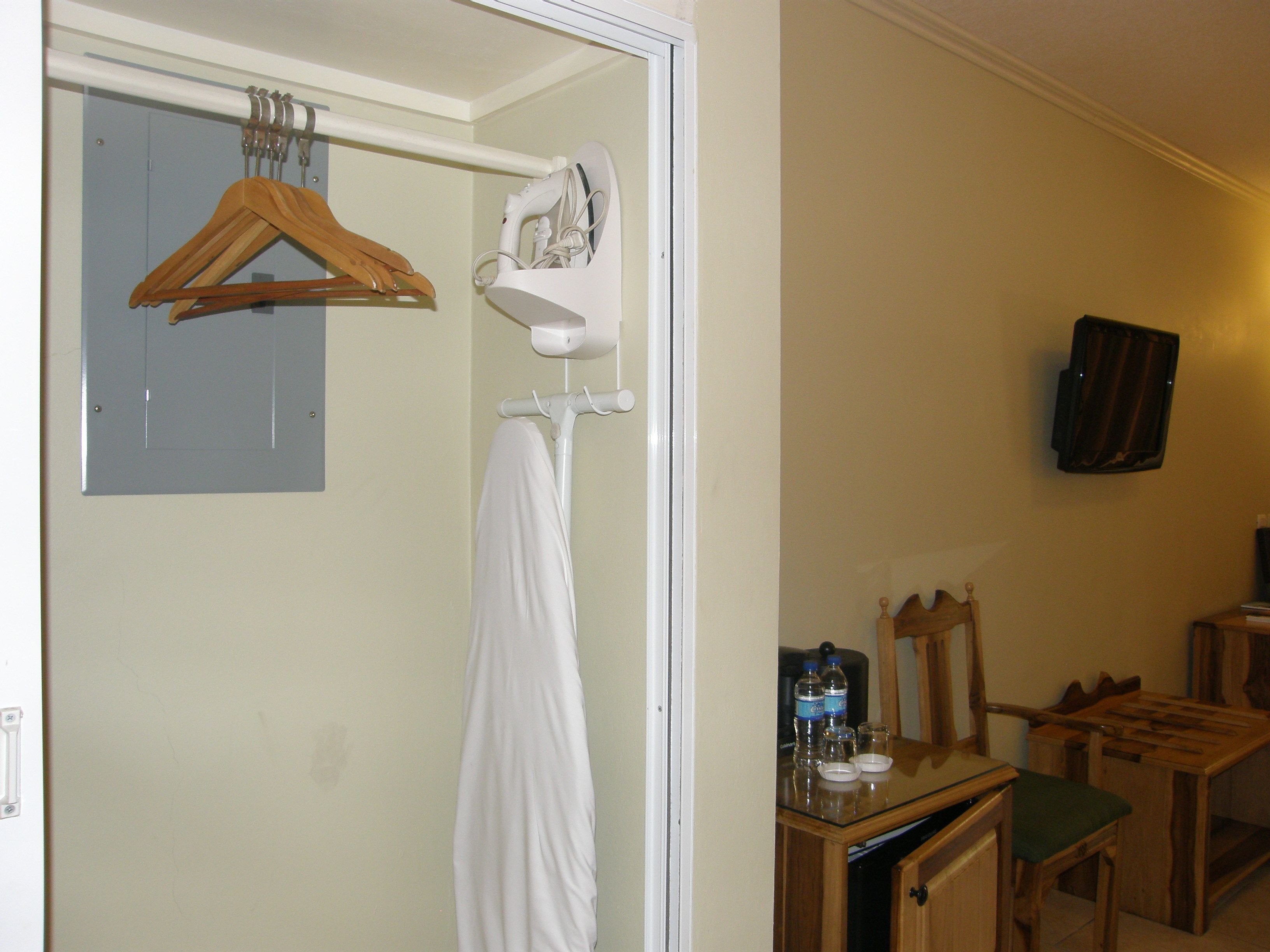 Une salle de bain avec une serviette suspendue à un porte-serviettes, un cintre en bois, une pomme de douche blanche et un miroir. On y trouve une petite table avec une bouteille d’eau et un verre.