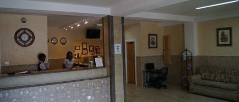 A reception area with a counter, two receptionists, a television, a couch, and a wall with clocks and framed pictures.