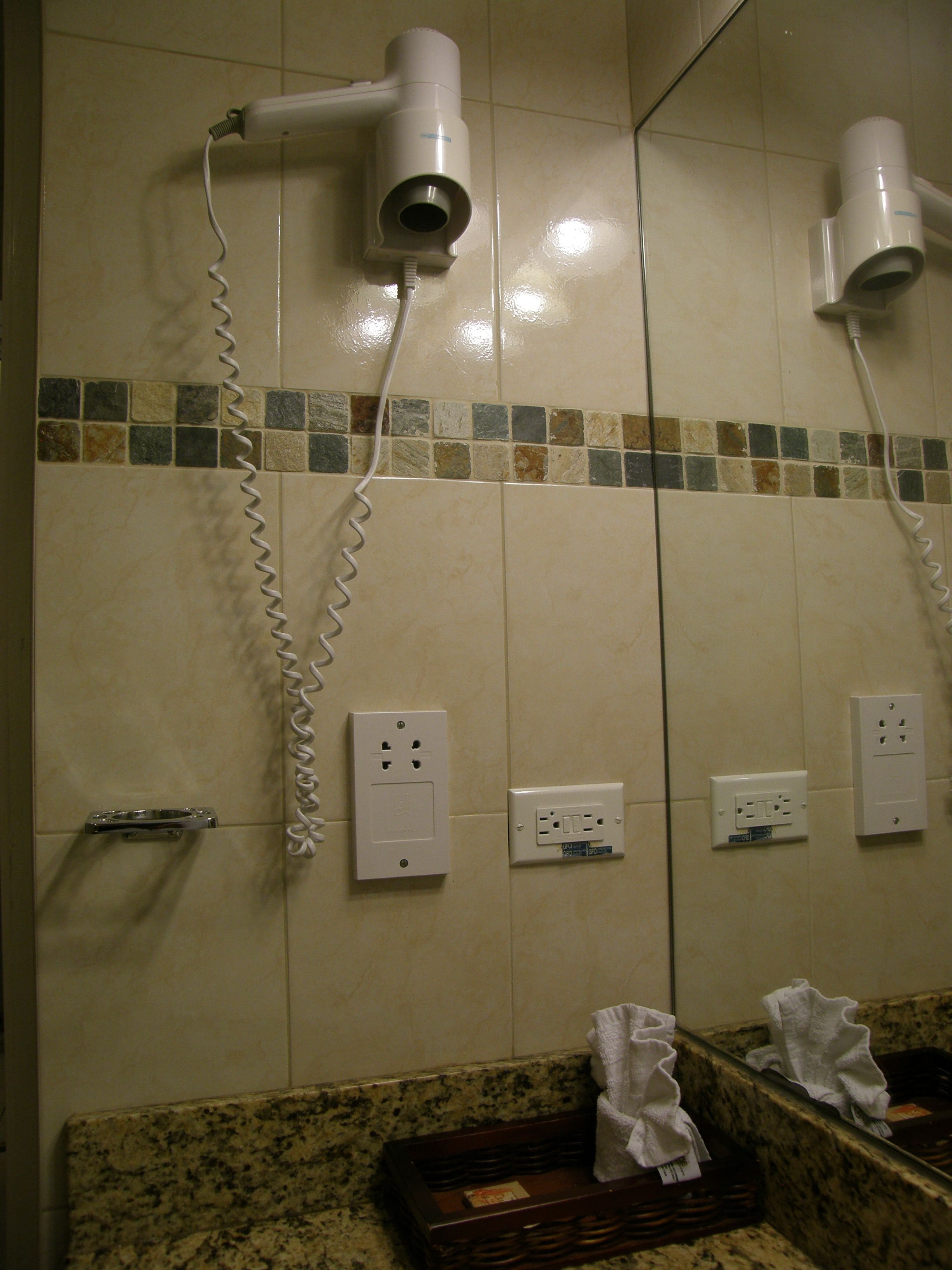 A bathroom with a hairdryer mounted on the wall, a mirror, and a countertop with a soap dish and a towel.