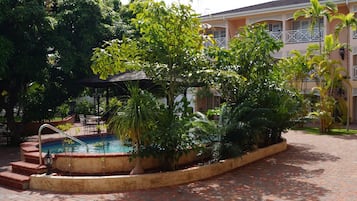 Un espace piscine agrémenté d’un chemin en briques, entouré d’une végétation luxuriante et bordé d’un bâtiment doté de balcons.