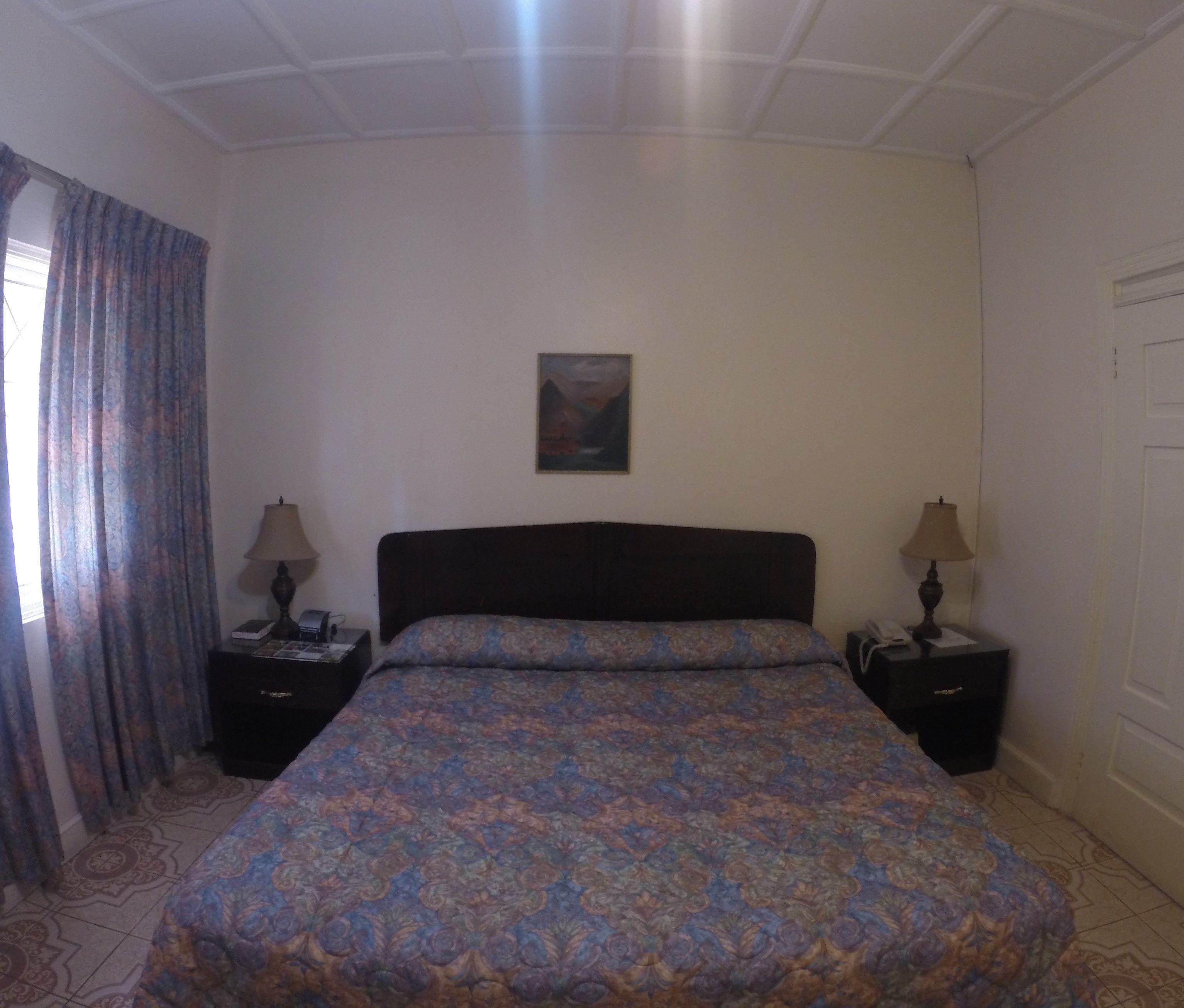 Une chambre d’hôtel avec un lit, deux tables de chevet avec des lampes, un tableau au mur et une fenêtre avec des rideaux.