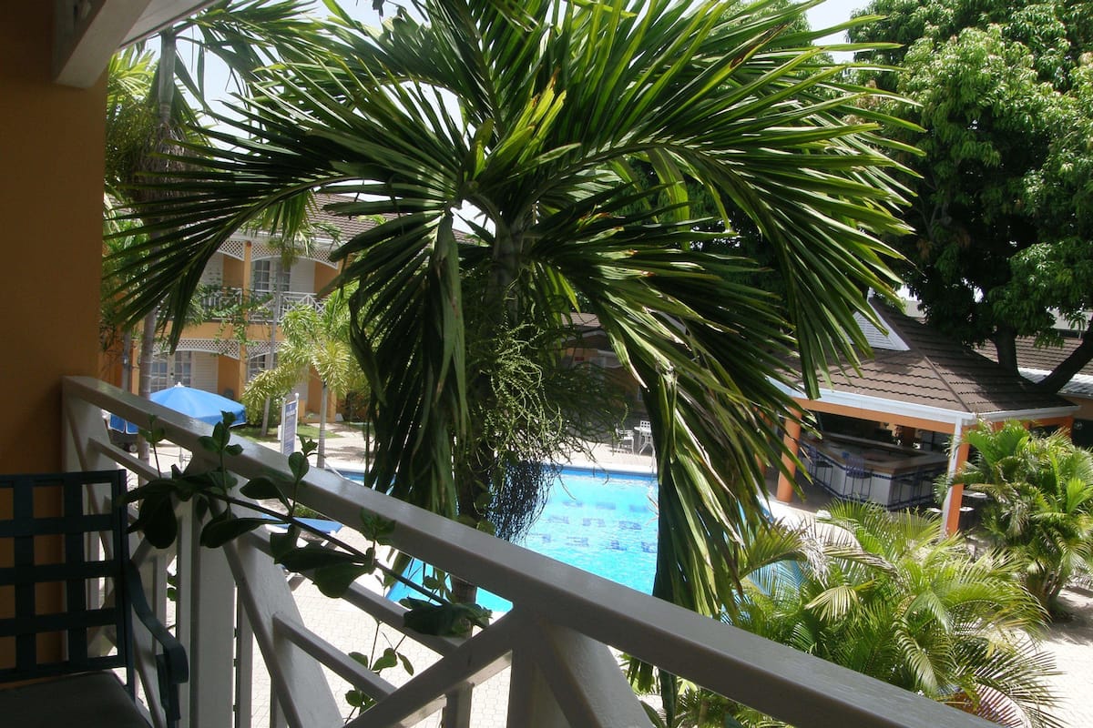 Un balcón con vista a una piscina rodeada de palmeras y un edificio con techo de tejas.