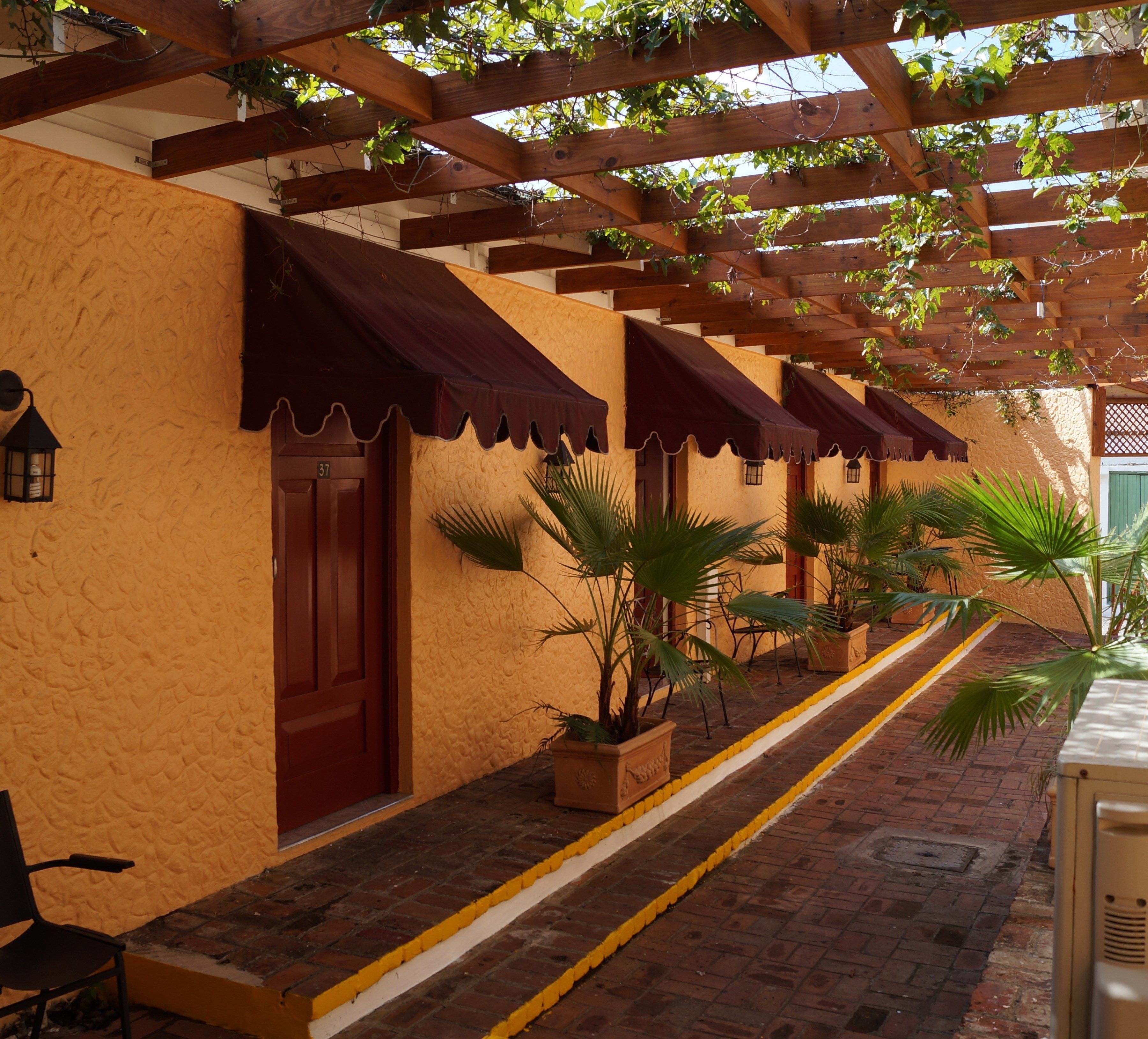 A covered outdoor area with wooden beams and a tiled floor, featuring a row of doors with awnings and potted plants.