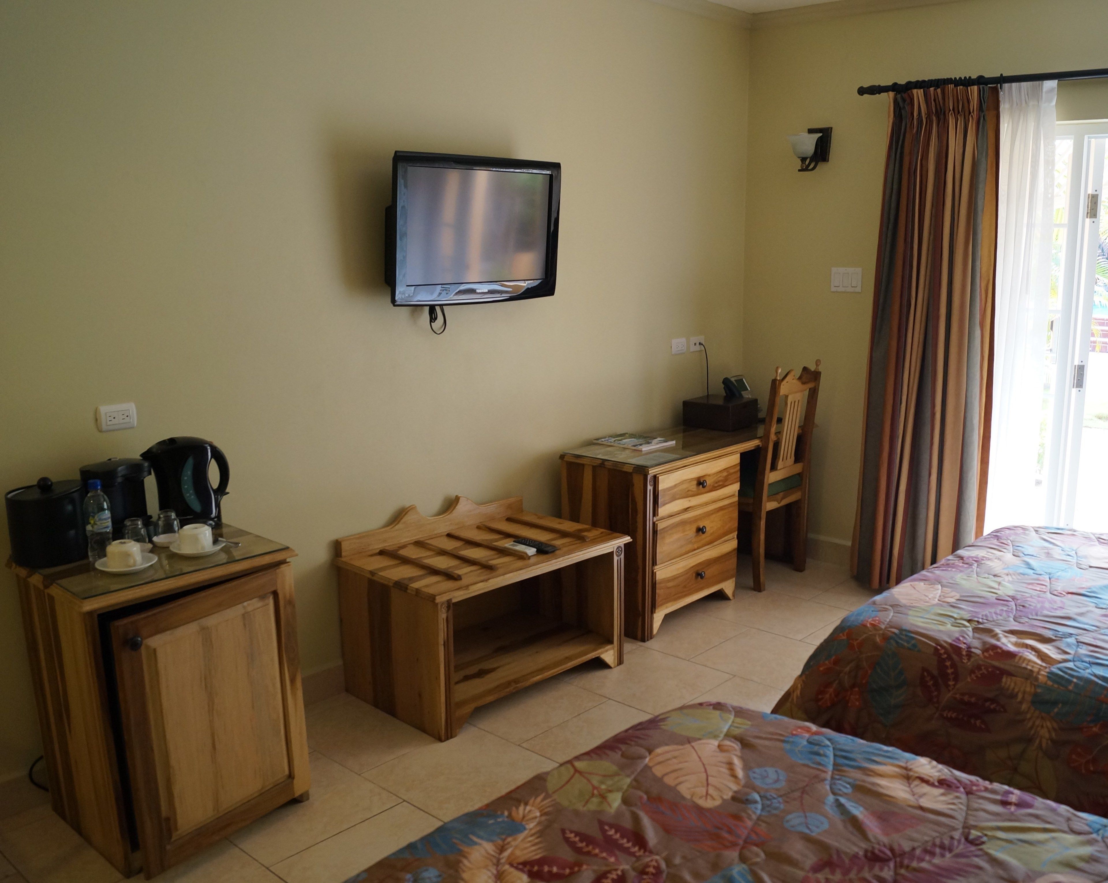 Habitación de hotel con cama, escritorio, silla, televisor, armario y ventana con cortinas.