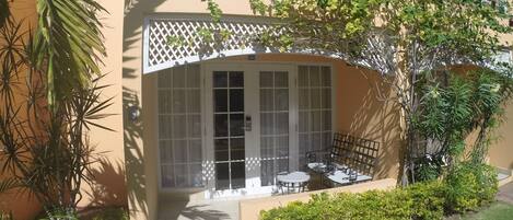A building with a covered patio area featuring a table and chairs, surrounded by greenery and a small garden.