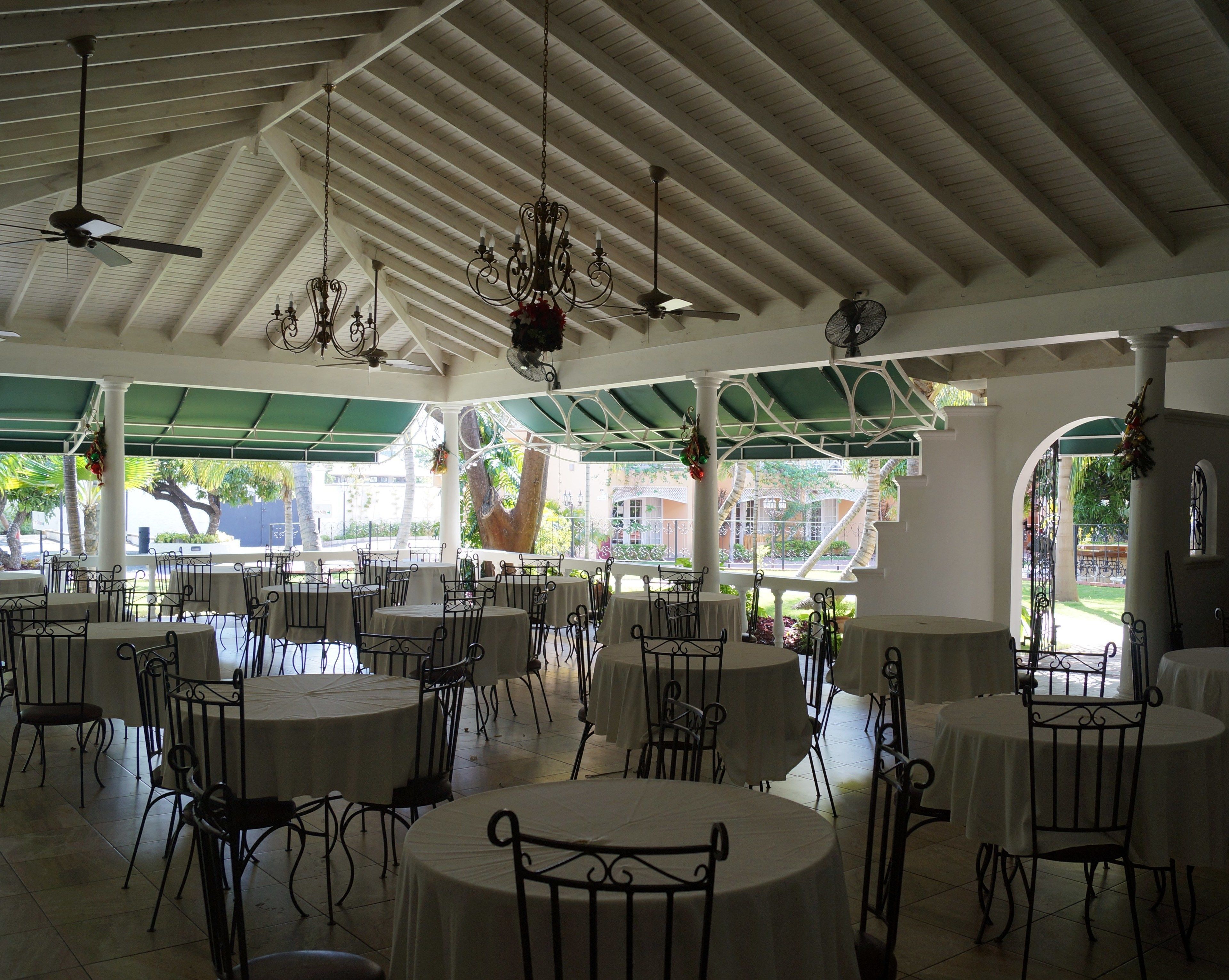 A spacious indoor area with multiple round tables and chairs, a high ceiling with wooden beams, and a view of greenery outside.