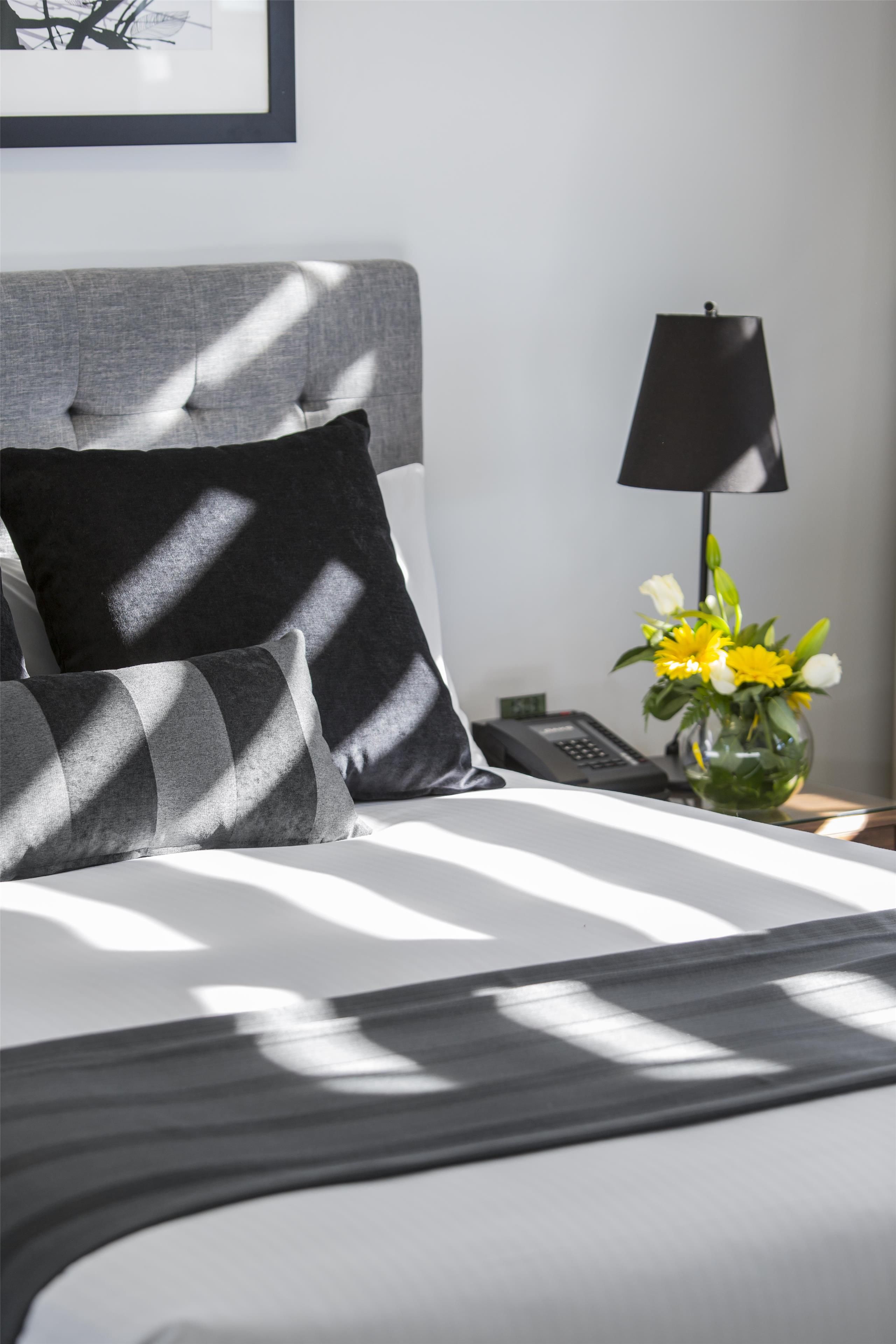 A neatly made bed with a grey headboard, a black and grey patterned pillow, a black lamp, and a vase with yellow flowers on a bedside table.