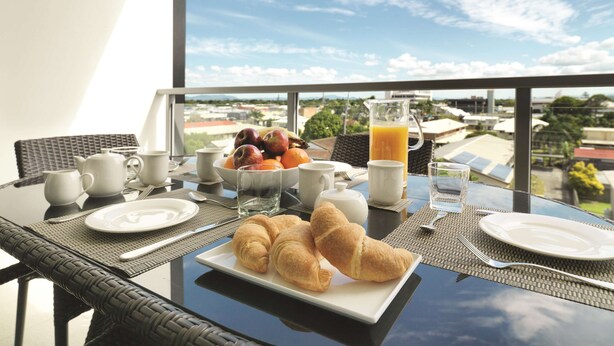 A breakfast table with croissants, fruit, and orange juice, overlooking a cityscape. - Oaks Mackay Carlyle Suites (Mackay)
