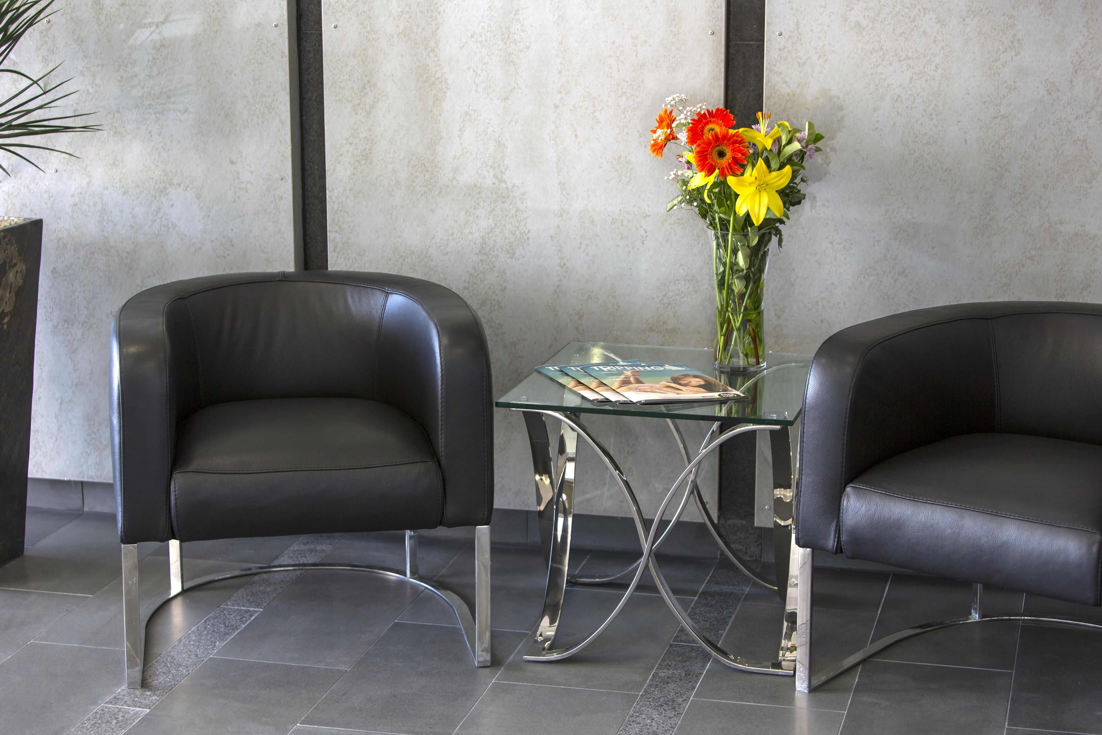 Two black leather chairs with metal legs, a glass table with a magazine, and a vase with flowers.