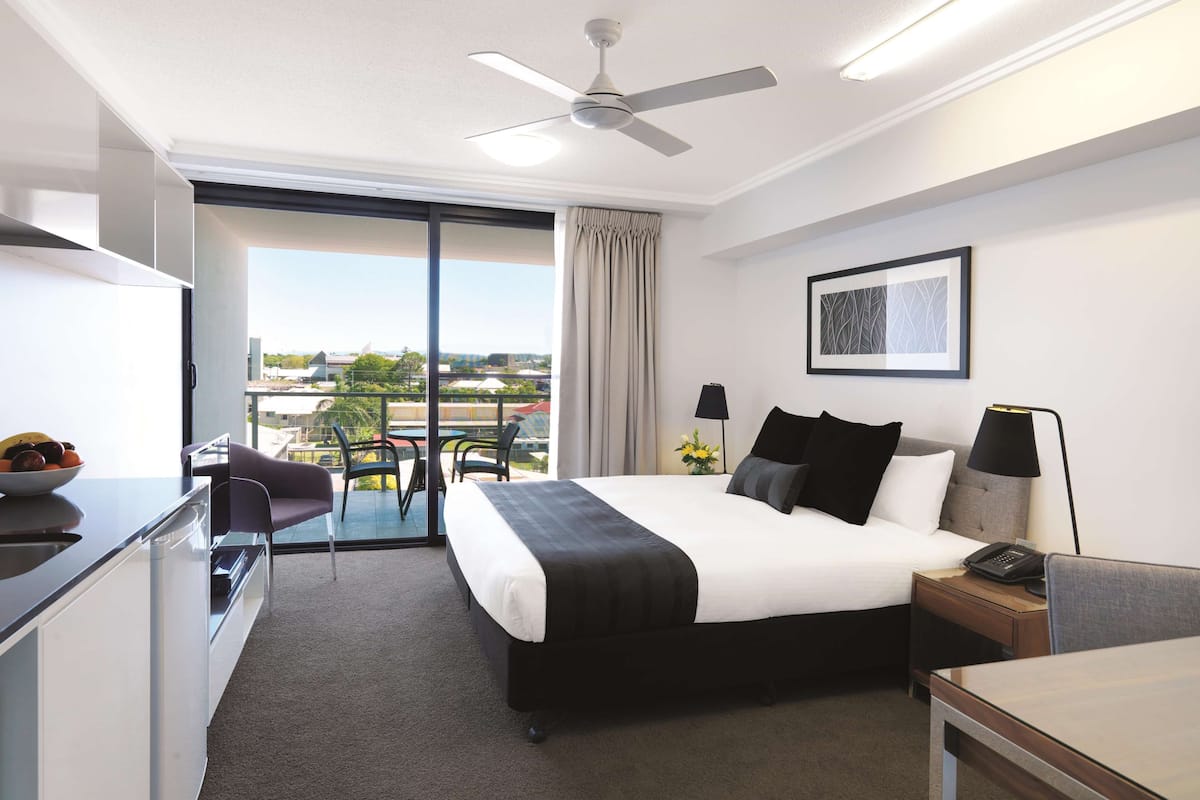 Ein Hotelzimmer mit Bett, Schreibtisch, Stuhl, Deckenventilator, einem Fenster mit Vorhängen, einem gerahmten Bild an der Wand und Blick auf die Stadt.