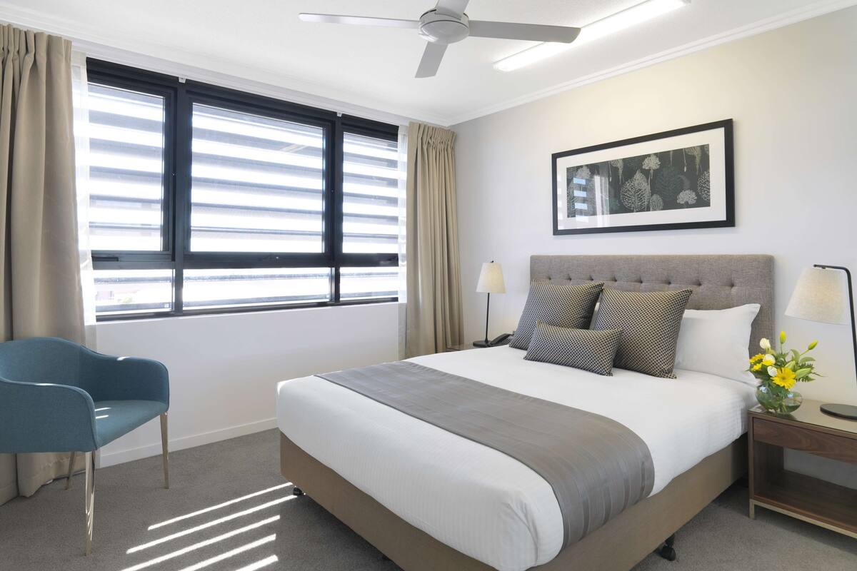 Ein Hotelzimmer mit einem Bett, einem Sessel, einem Deckenventilator, einem Fenster mit Jalousien, einem gerahmten Bild an der Wand und einem Nachttisch mit Lampe und Blumen.