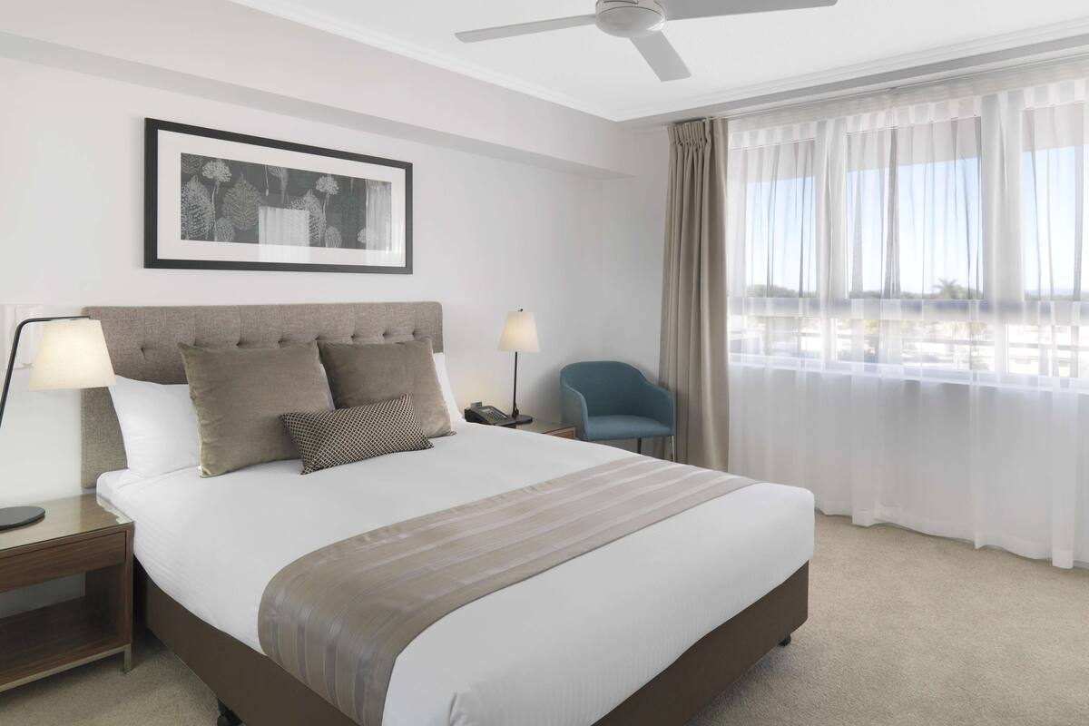 Ein Hotelzimmer mit einem Bett, einem Deckenventilator, einem gerahmten Bild an der Wand, einem Sessel und einem Fenster mit Vorhängen.