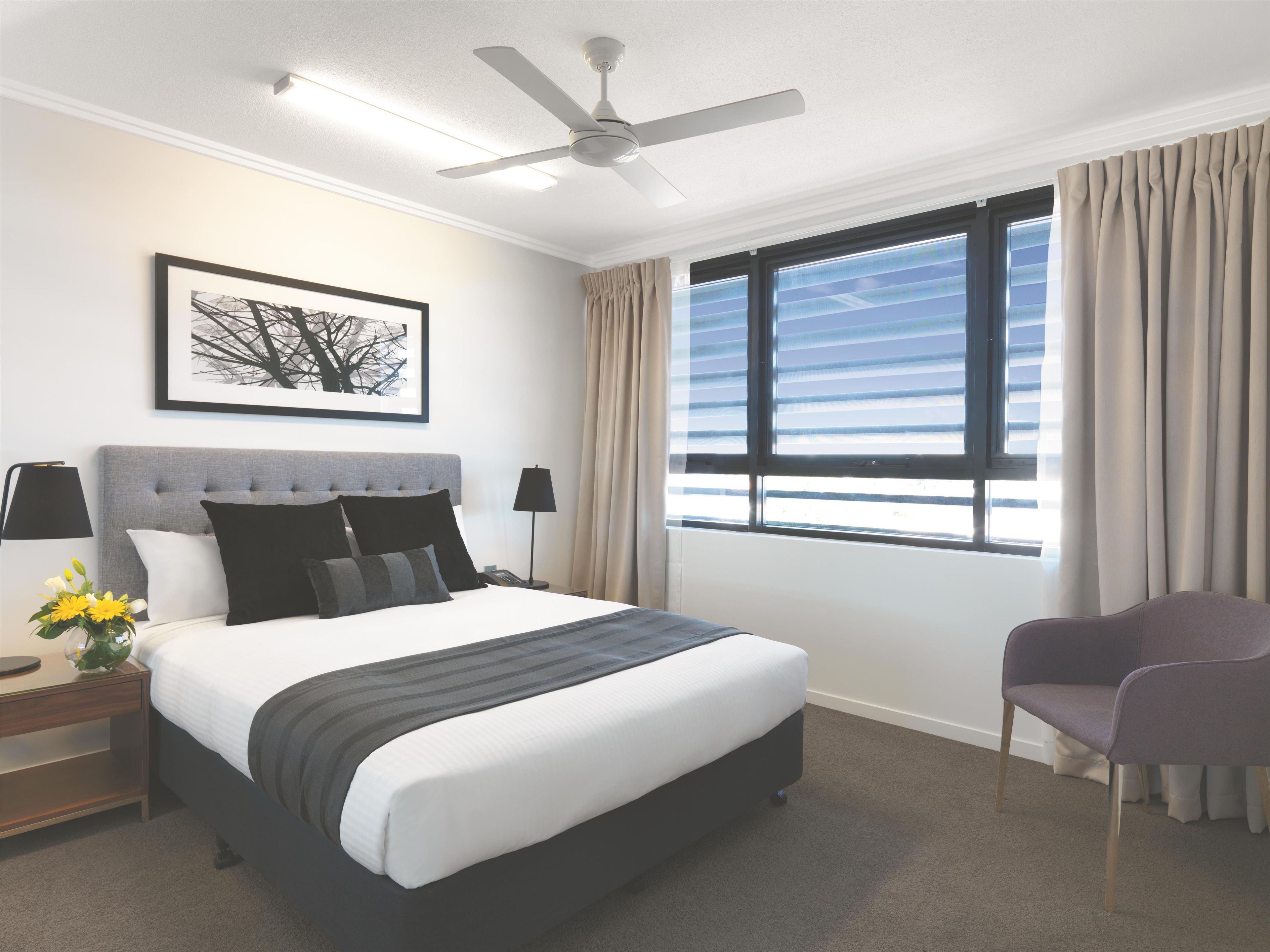 Et hotelværelse med seng, stol, ventilator, et vindue med gardiner, et sengebord med lampe og et billede i ramme på væggen.