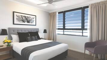 Et hotelværelse med seng, stol, ventilator, et vindue med gardiner, et sengebord med lampe og et billede i ramme på væggen.