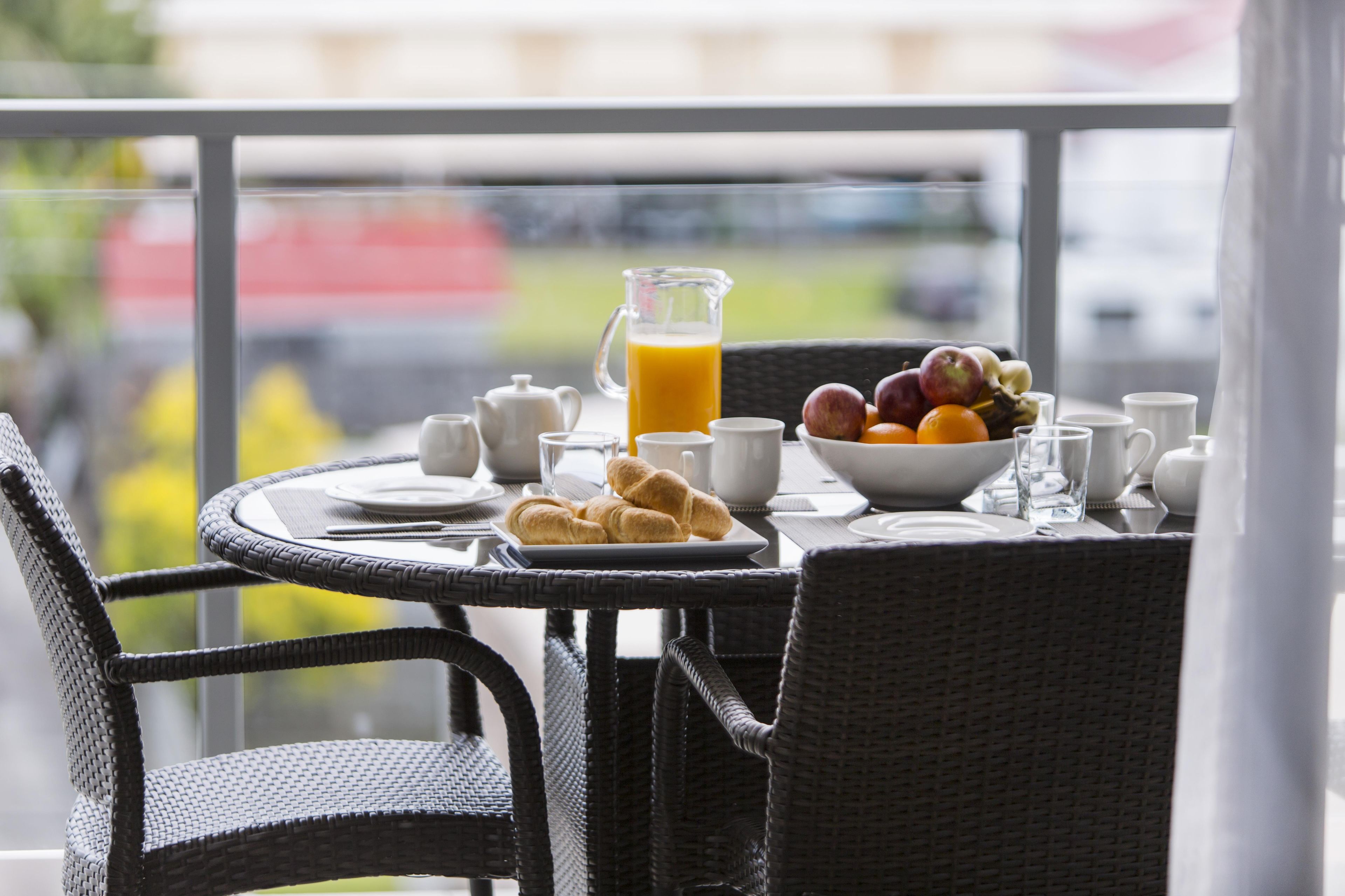 Una mesa de desayuno con cruasanes, un frutero, una jarra de jugo de naranja y varias tazas y platitos.
