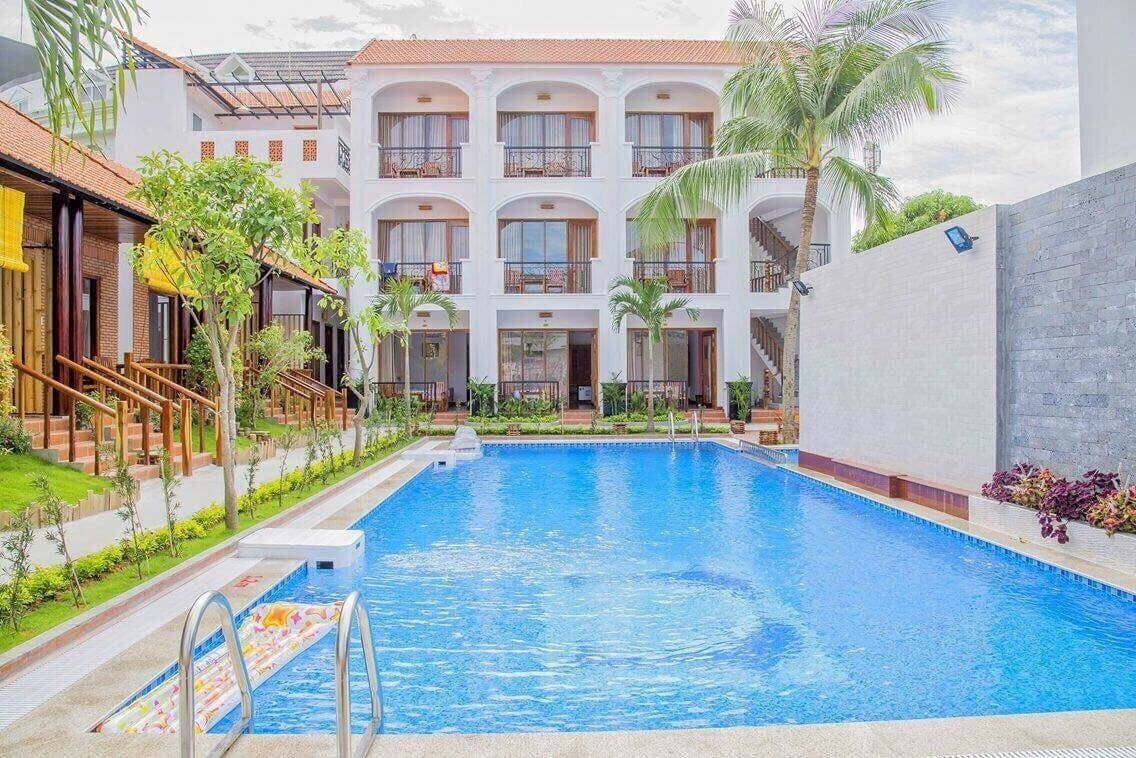 A swimming pool with a clear blue water, surrounded by a white building with balconies and palm trees.